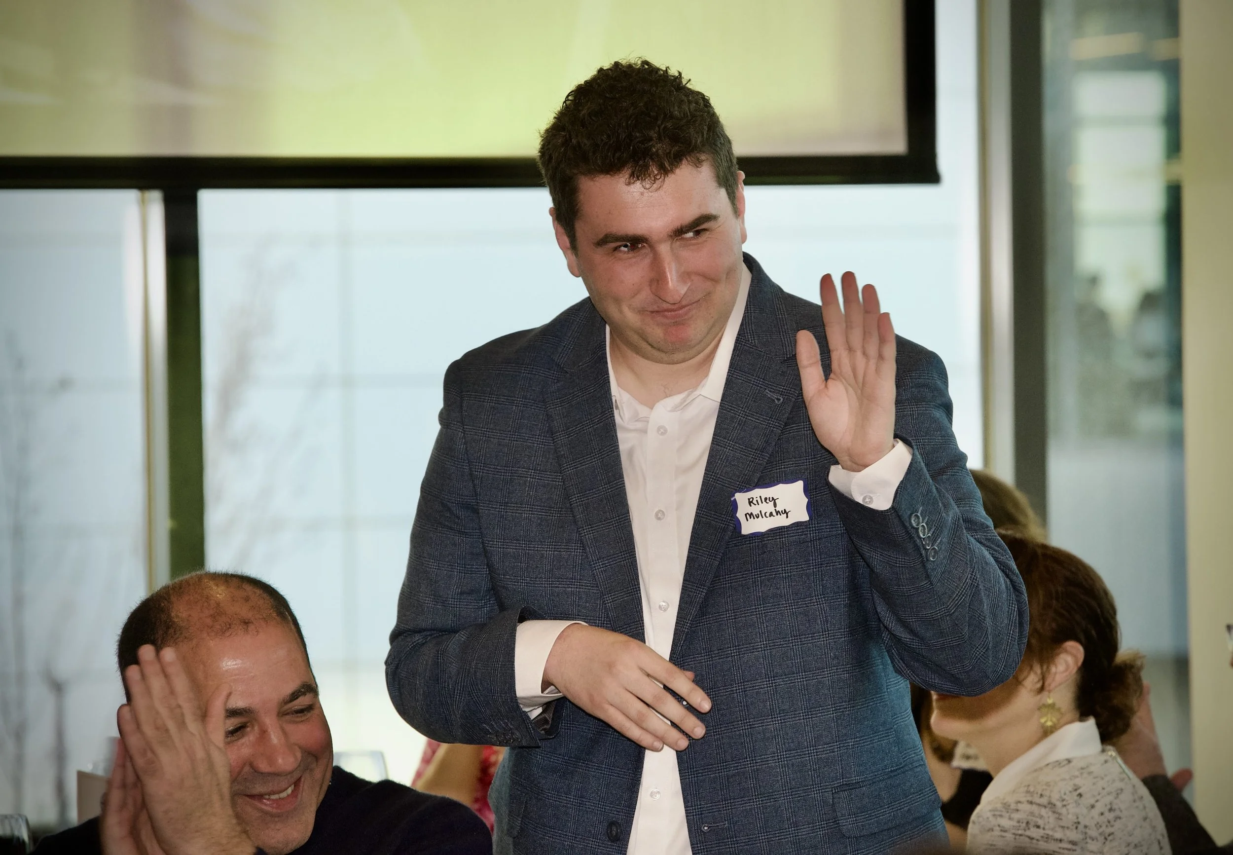 A man in a suit with a name tag that reads 'Riley Mulcahy' is standing and smiling, raising his hand in a wave during a meeting or conference. Other people are seated around him, engaged and smiling.