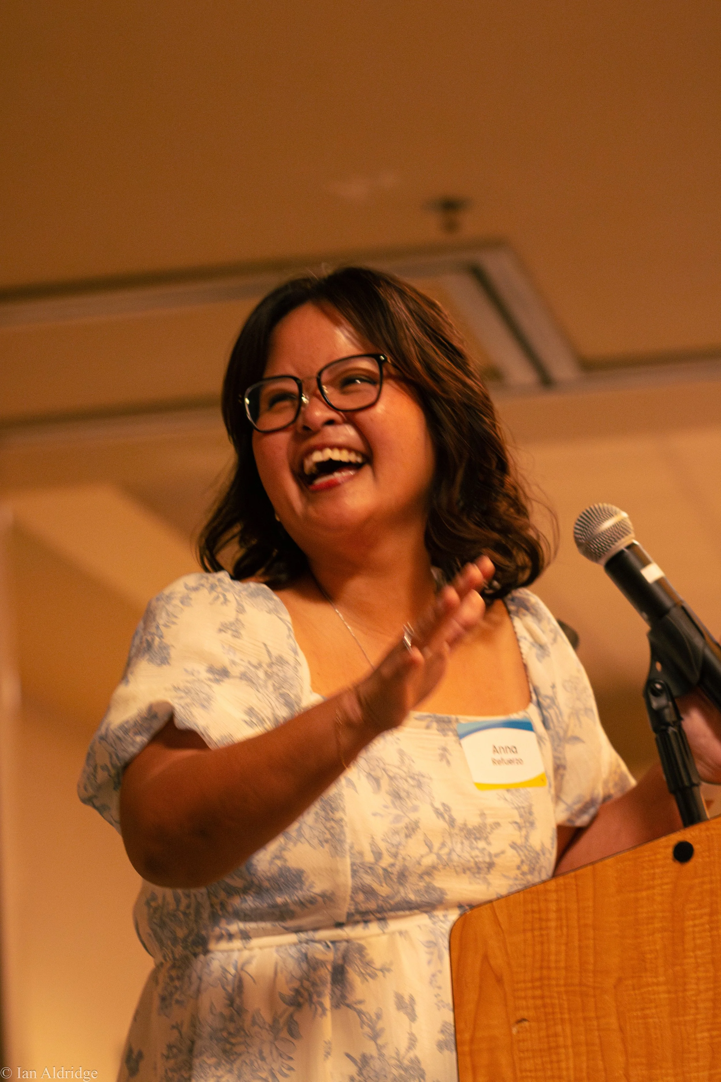 A woman with glasses, wearing a white and blue patterned dress, laughing and speaking into a microphone at a gathering or event.