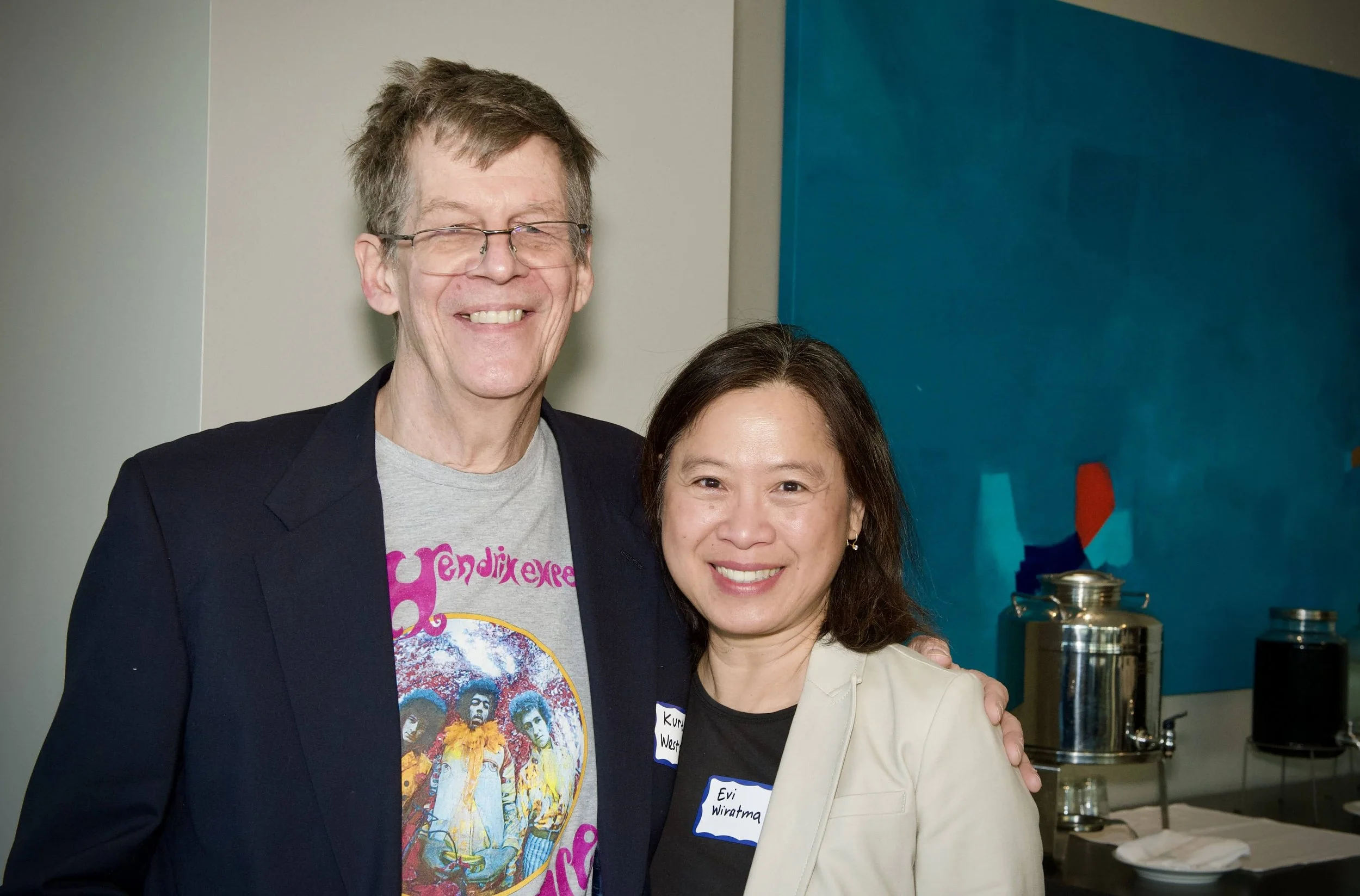 Two smiling women posing together indoors, one man standing behind them, near a blue wall and a table with a large metal water dispenser.