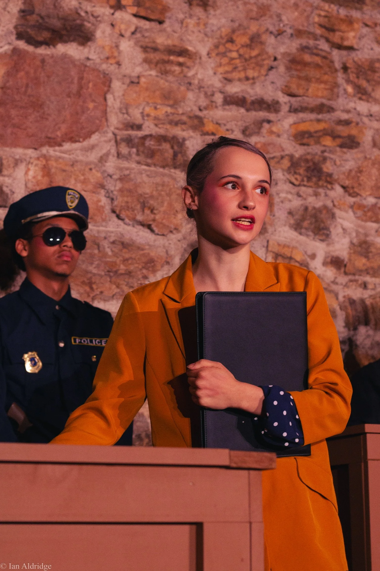 A woman with short hair and wearing an orange suit is holding a black folder and standing in front of a brick wall. A man in police uniform is seen in the background wearing sunglasses and a police hat.