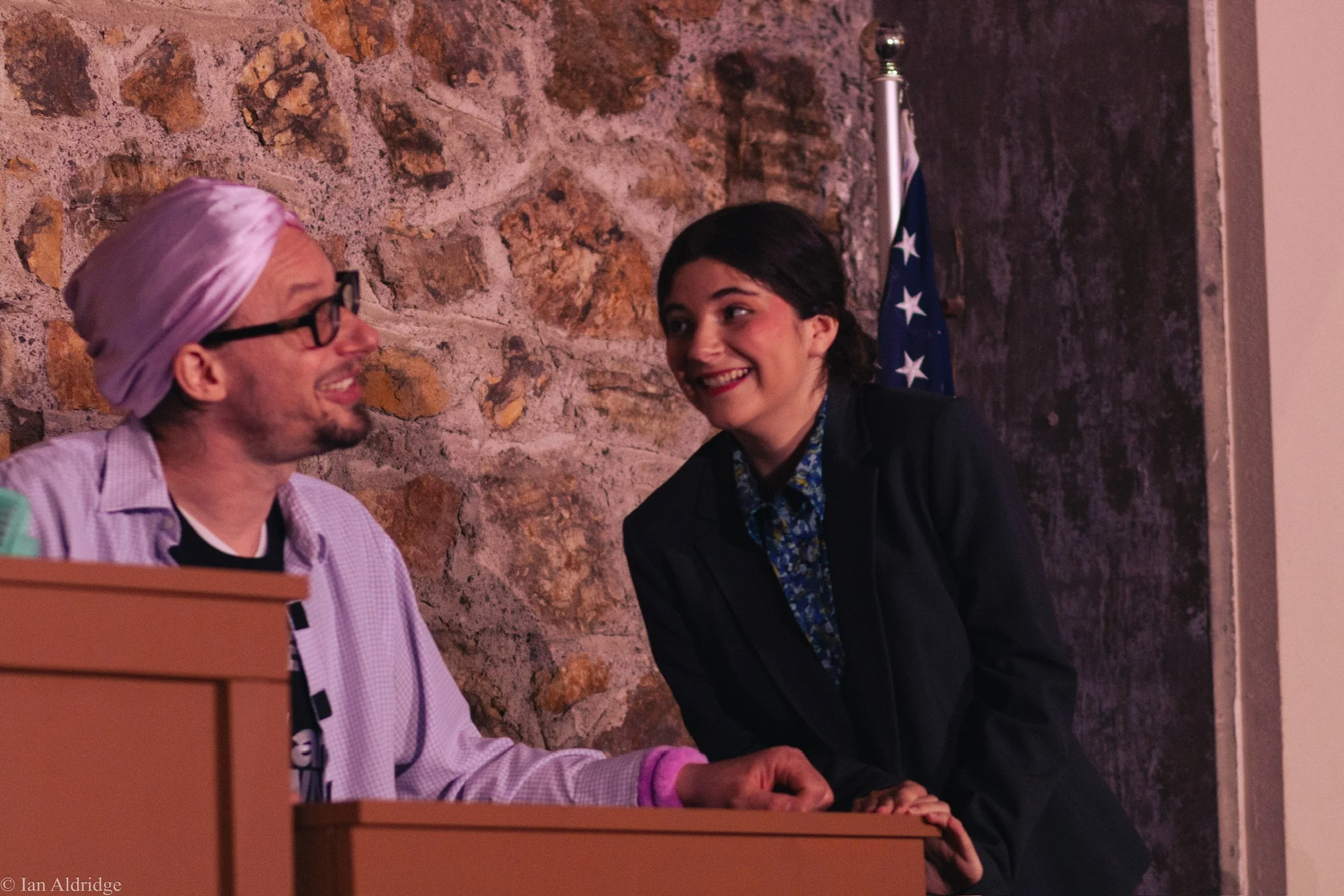 A man with glasses and a pink head covering smiling and talking to a woman with dark hair, wearing a black blazer, who is also smiling. They are in a room with a stone wall and an American flag in the background.