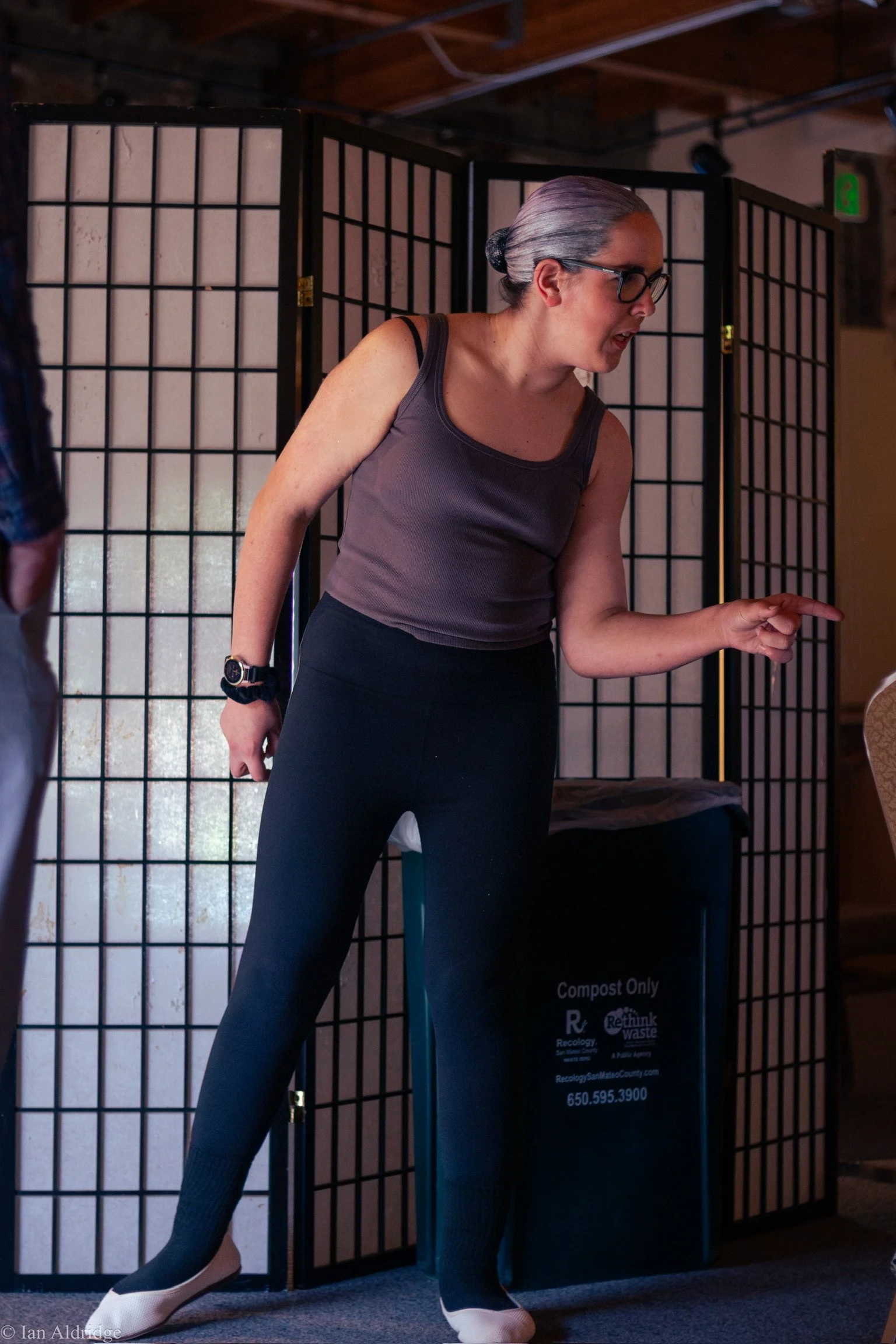 A woman with gray hair tied back, wearing glasses, a sleeveless top, and leggings, standing in front of a black folding screen. She appears to be talking or explaining something.
