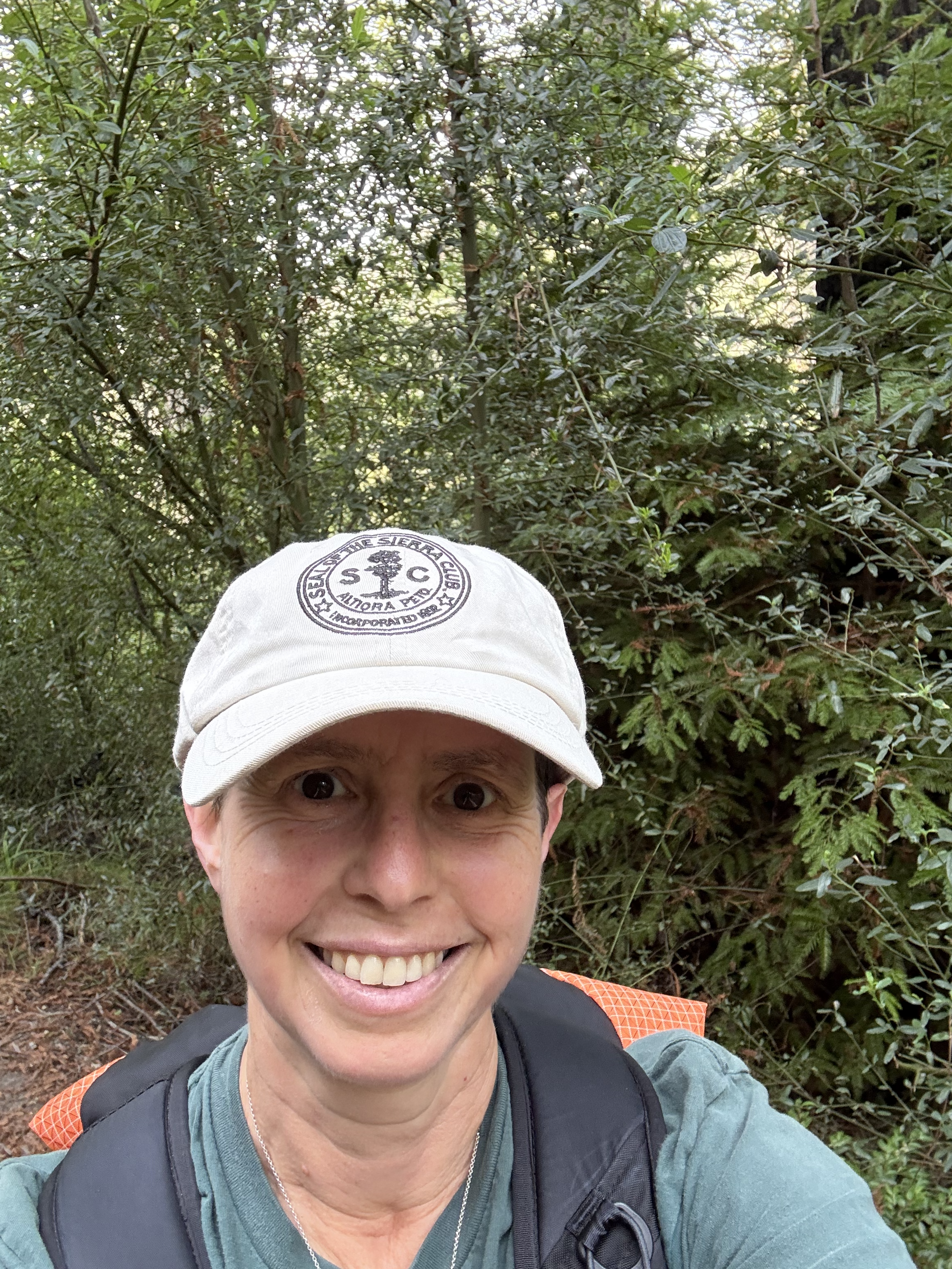 A person smiling while hiking in a forest with green foliage, wearing a white cap, gray shirt, and backpack.