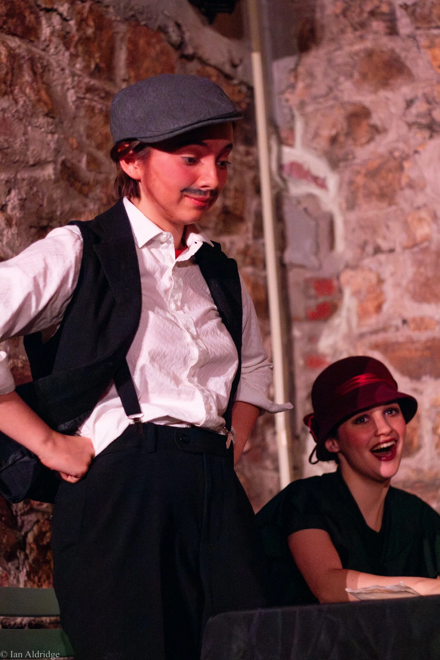 A person wearing a gray newsboy cap, a white shirt, black vest, and black pants stands against a brick wall, smiling slightly and looking downward. Another woman wearing a black hat with red accents and a black top, smiling and laughing, sits below h
