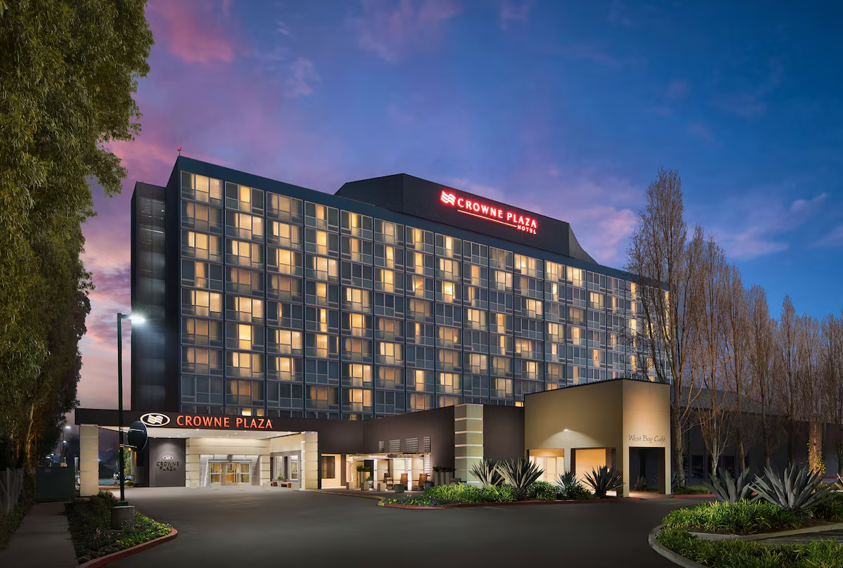 Nighttime exterior view of Crowne Plaza hotel with illuminated windows, front entrance, and surrounding landscaping, including trees and desert plants.