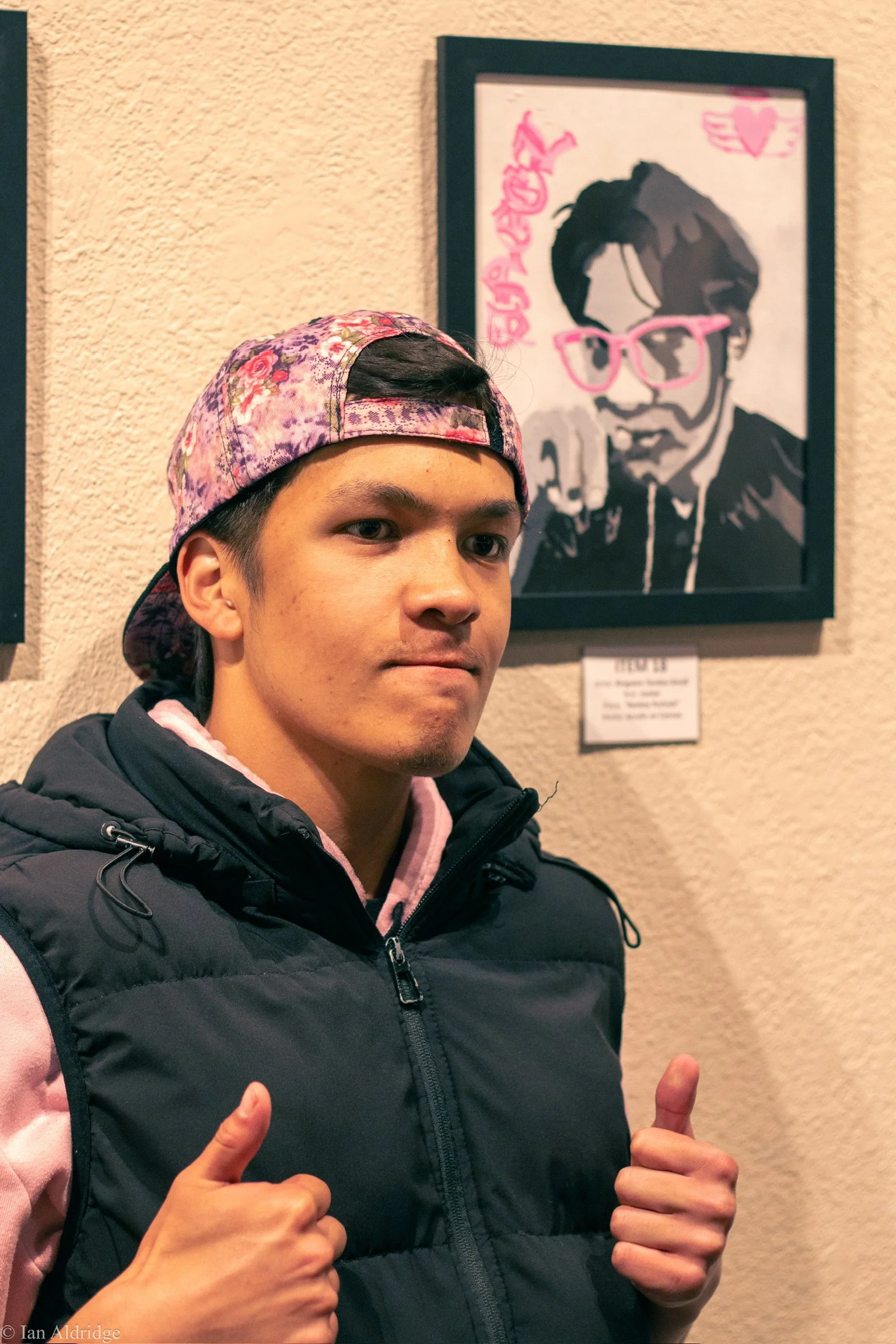 A young man wearing a pink and purple floral baseball cap and a dark quilted vest giving two thumbs up. He is standing in front of a framed art piece of a person with sunglasses and headphones, with some pink graffiti-style writing nearby.