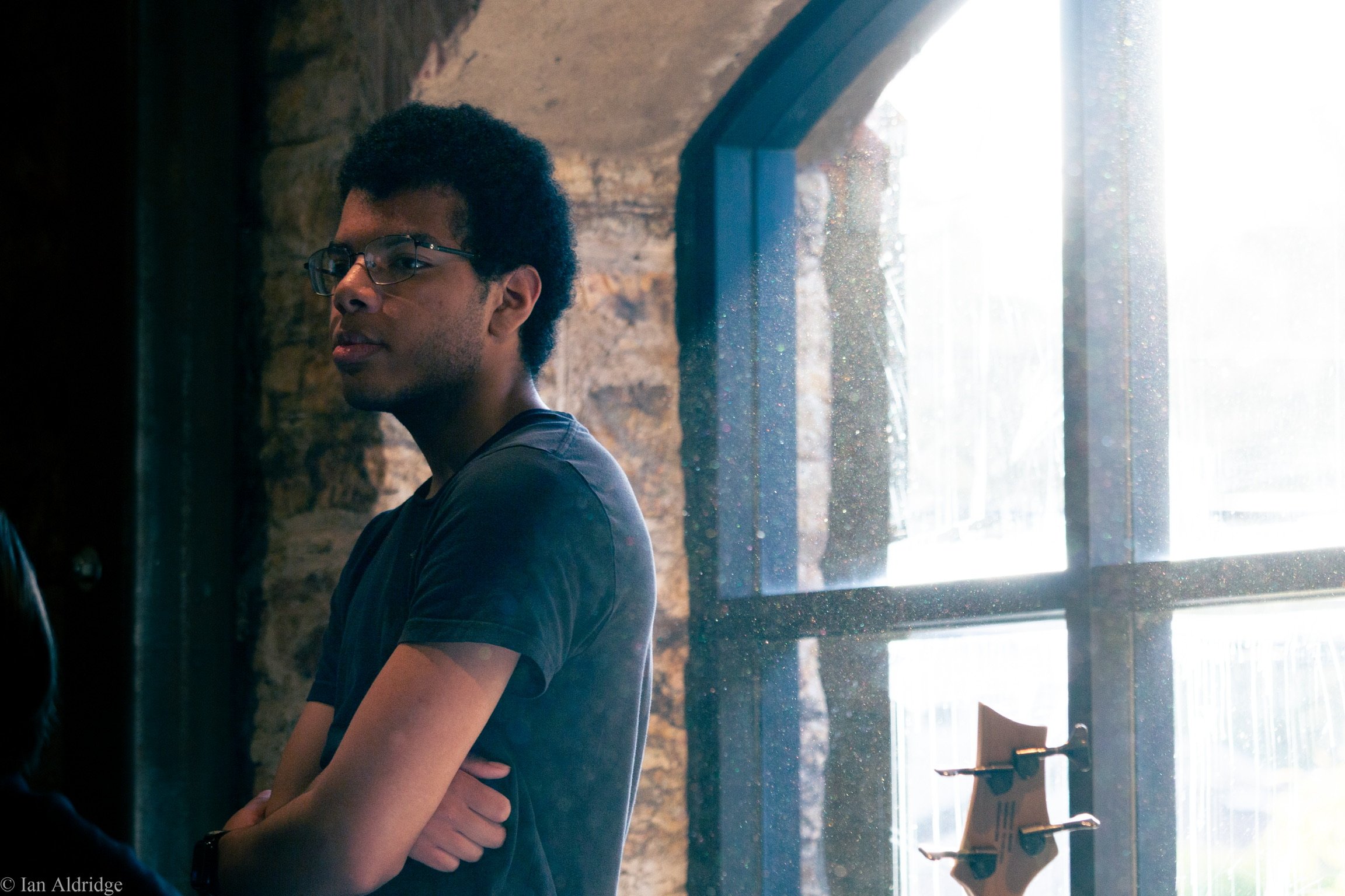A young man with glasses and curly black hair, standing indoors near a window with a brick wall behind him. He is wearing a dark t-shirt and looking towards the camera with a neutral expression.