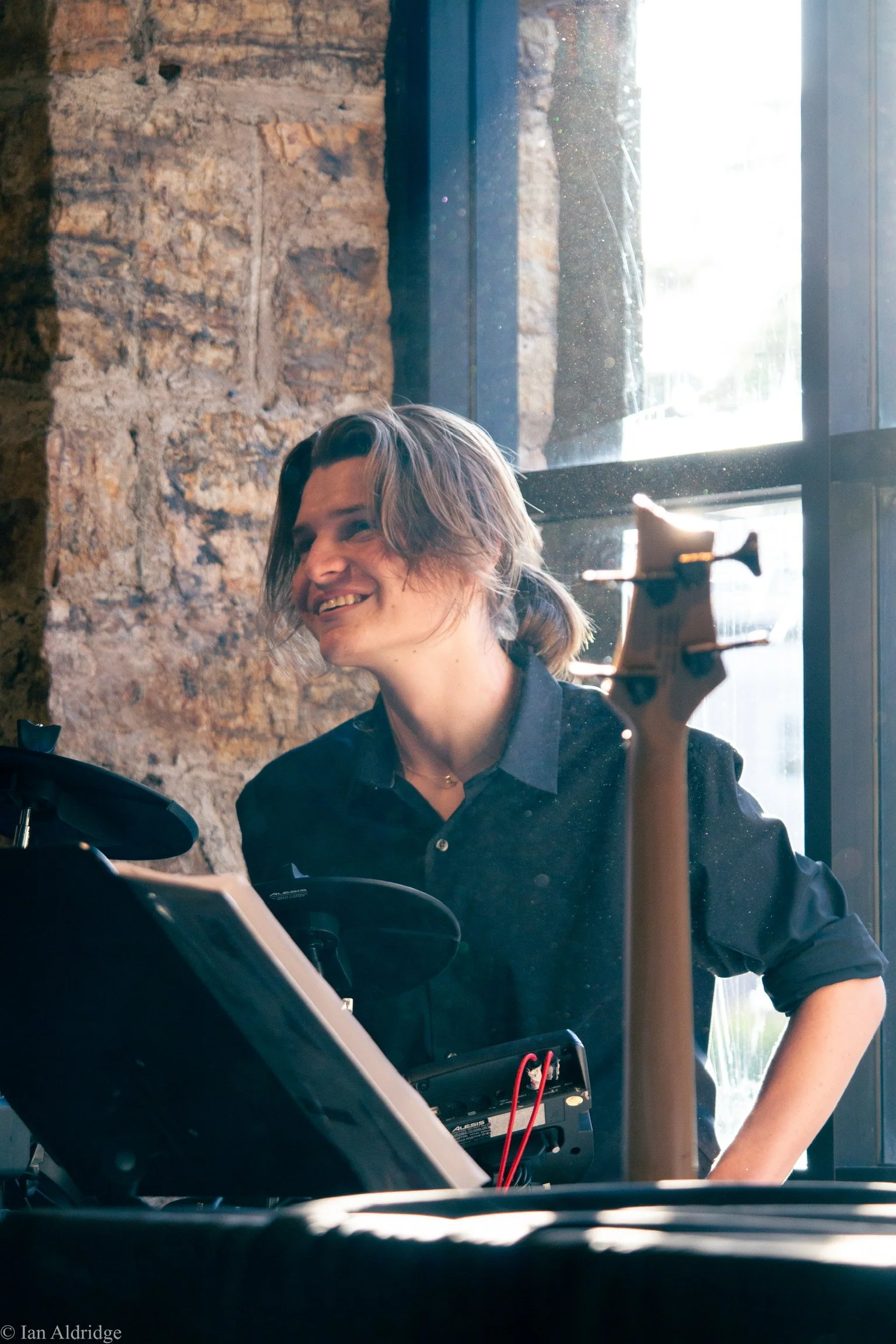 Person with shoulder-length hair and a black shirt smiling inside a building near a window, with a brick wall in the background.