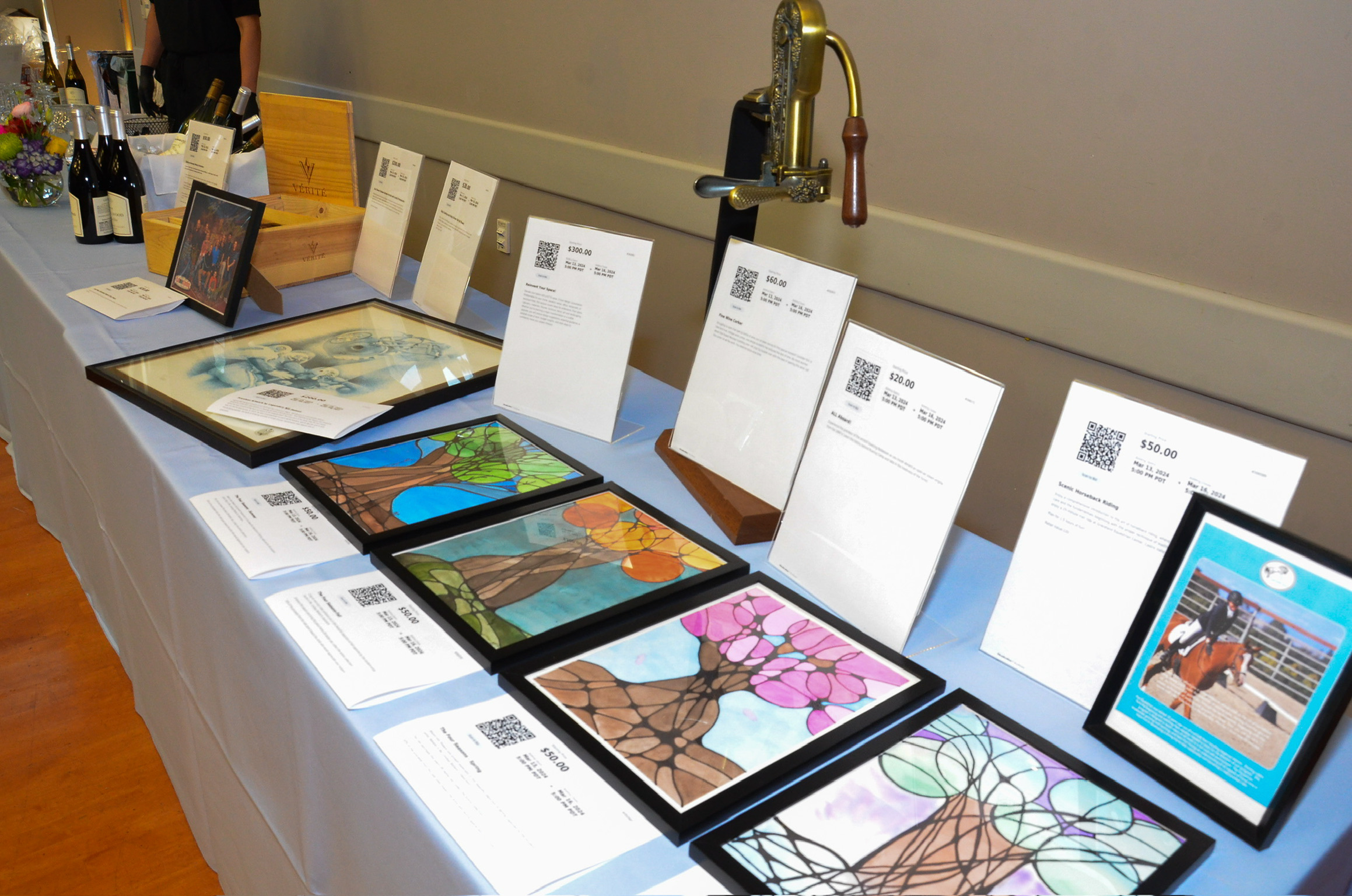 A table displaying various pieces of artwork, mostly stained glass designs, with informational placards and prices. In the background, bottles of wine and a person wearing black gloves are visible.