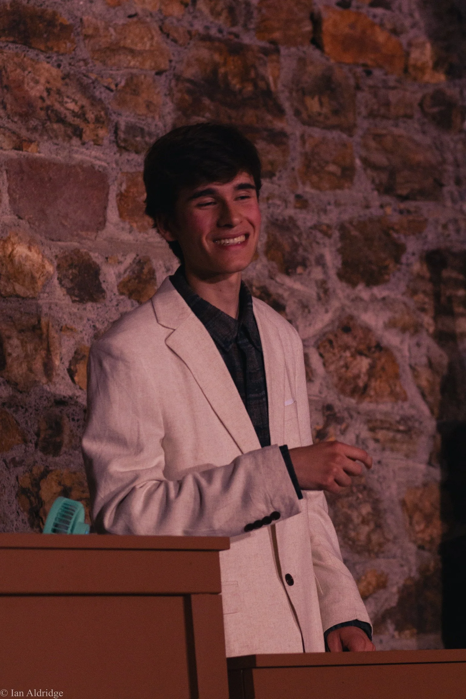 A young man with dark hair, wearing a beige blazer over a dark plaid shirt, standing against a brick wall. He is smiling with his eyes partly closed.