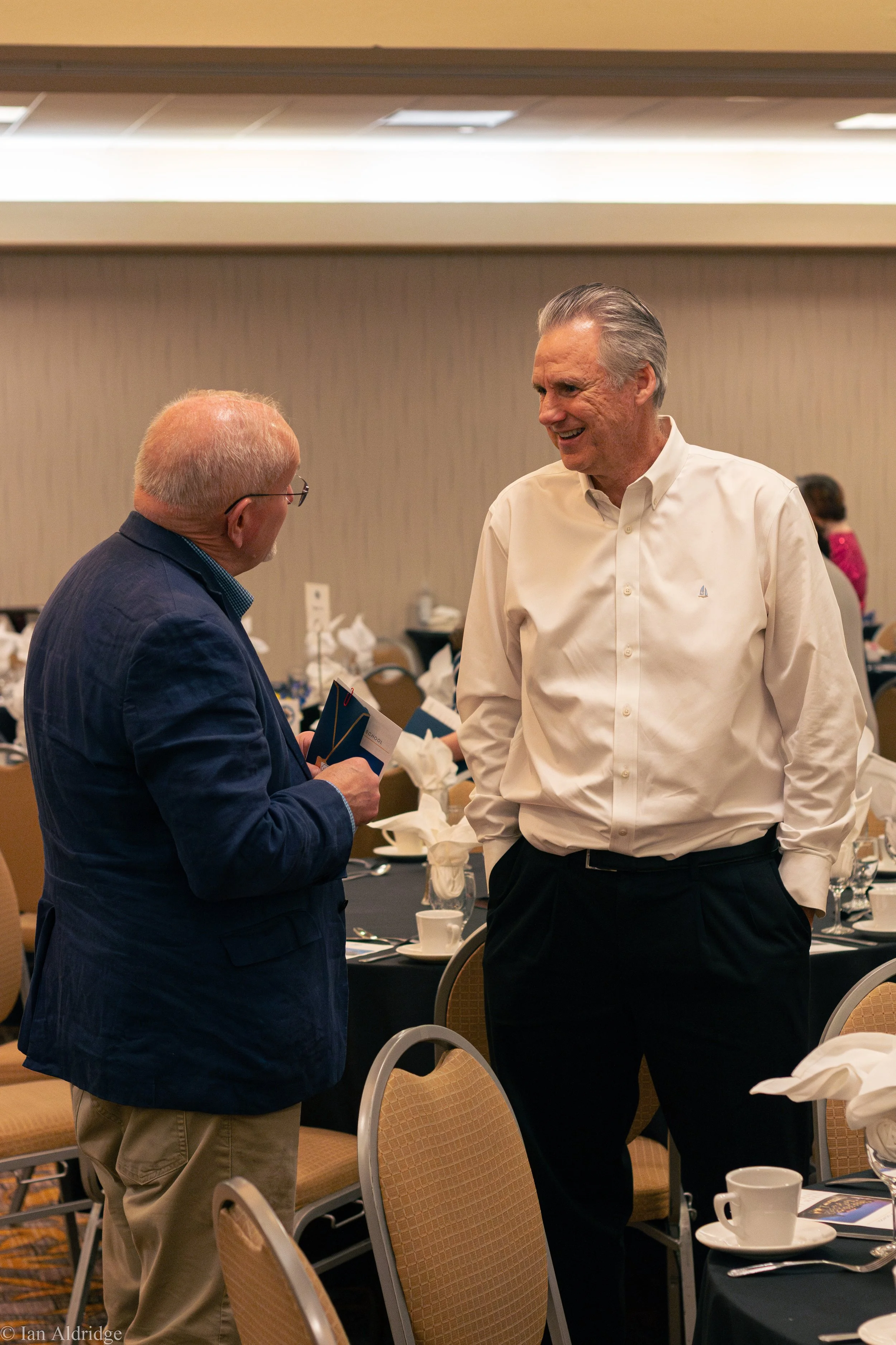 Two elderly men are engaged in conversation at a formal event. One man is wearing a navy blazer and beige pants, holding some papers, while the other man is dressed in a white button-down shirt and black pants. They are standing next to a table set w