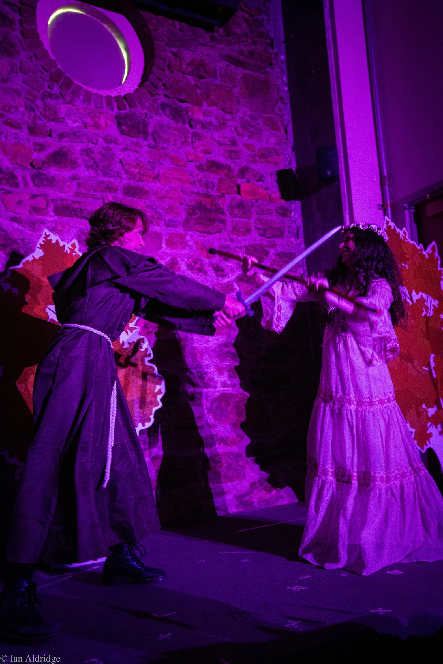 Two women engaged in a staged sword fight operation with foam swords, on a theatrical stage illuminated with purple lighting, against a brick wall backdrop with autumn leaves decoration.