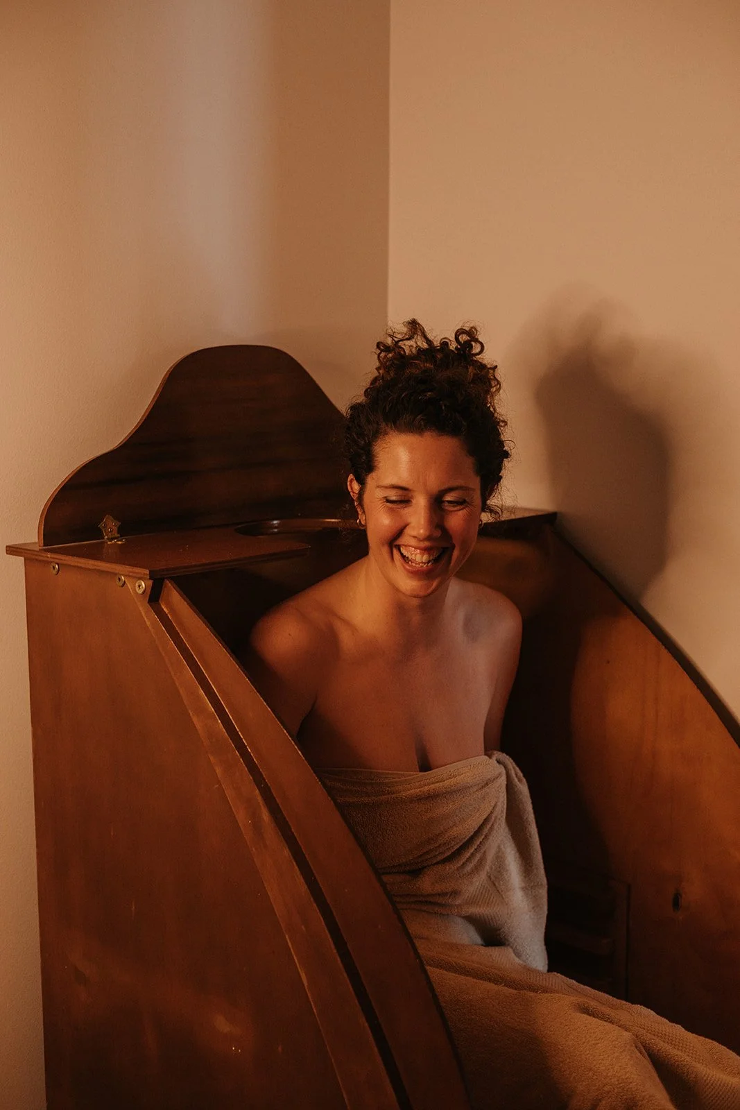 A woman wrapped in a towel, laughing and sitting in a wooden steam box, with curly hair and a light brown wall in the background.