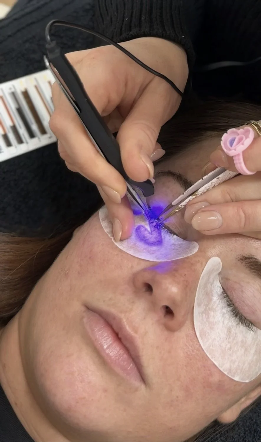A person undergoing a cosmetic eyelash enhancement procedure with a technician using specialized tools and UV LED light to cure the glue.