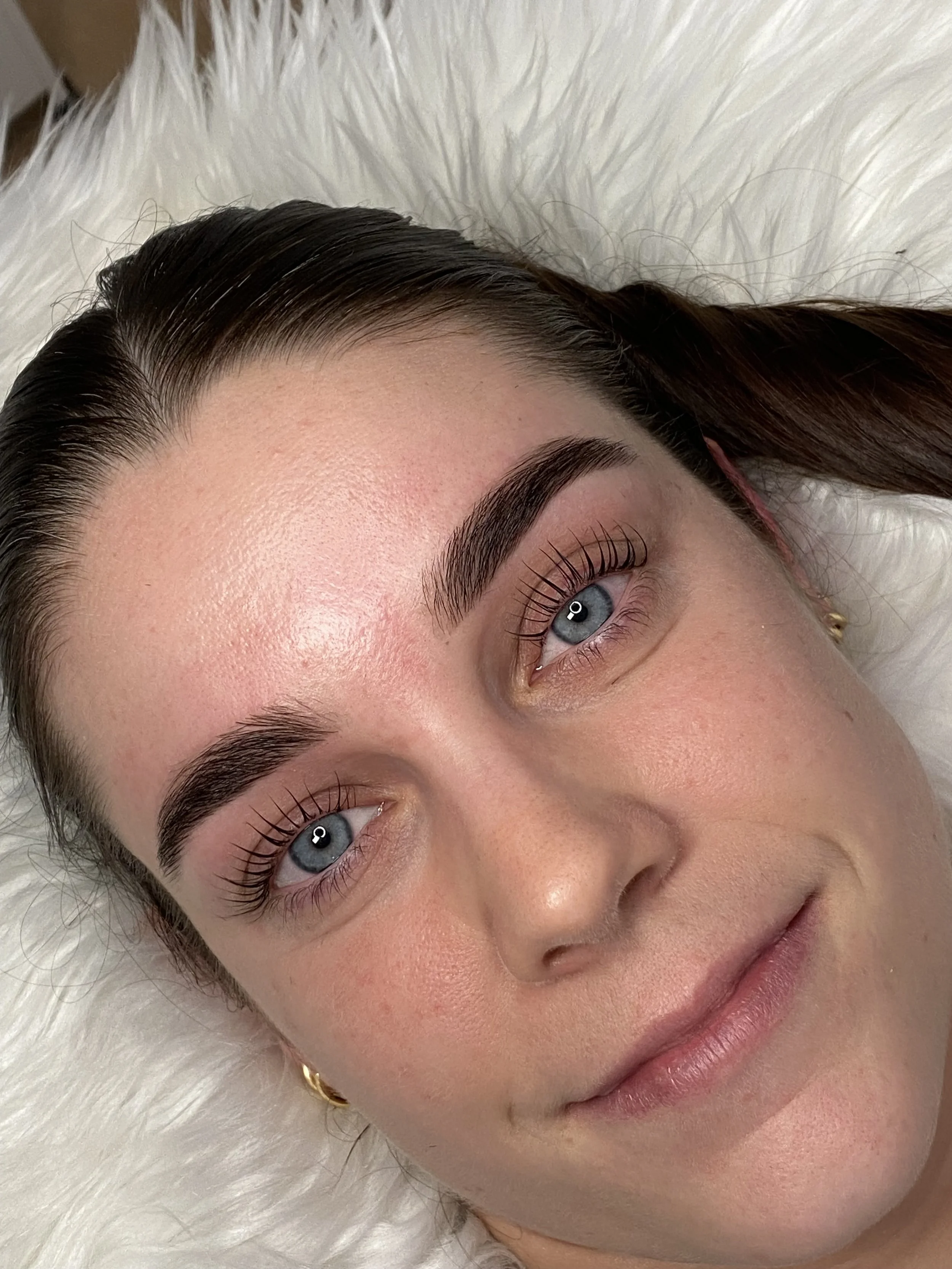 Close-up of a woman's face with well-groomed eyebrows, blue eyes, and smooth skin, lying on a white fluffy surface.