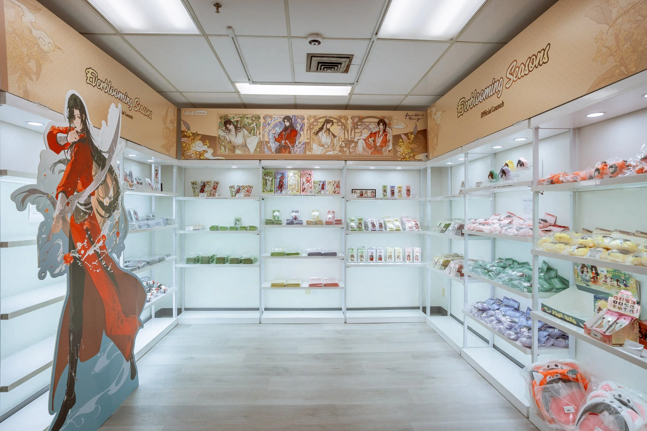 Store display with shelves filled with anime merchandise, including plush toys and figures, featuring characters in traditional Chinese or Japanese attire. Decorative banners with illustrations of characters are above the shelves.