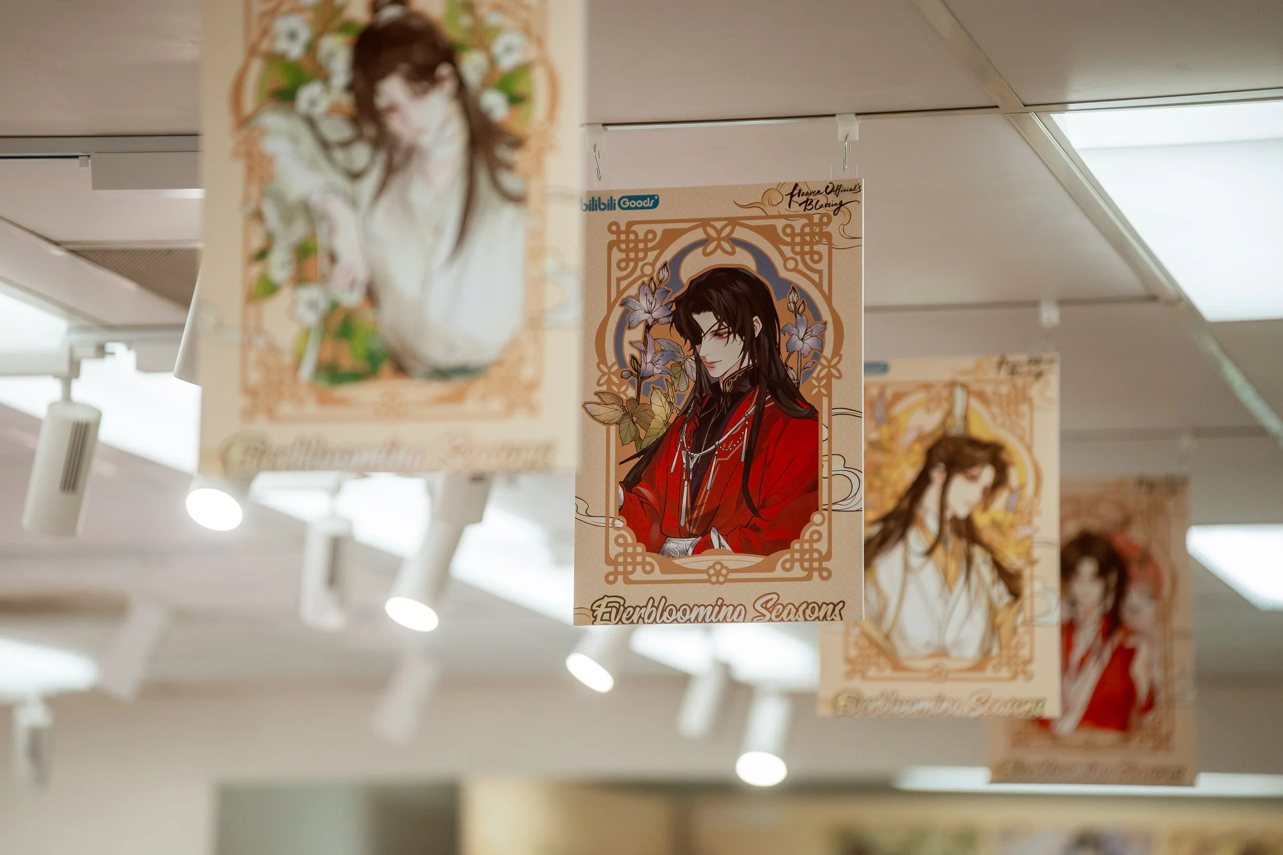 Banners featuring anime-style male characters with dark hair, one with red clothing and glasses, hanging from the ceiling of a store. The banners are associated with the theme 'Everblooming Seasons.'