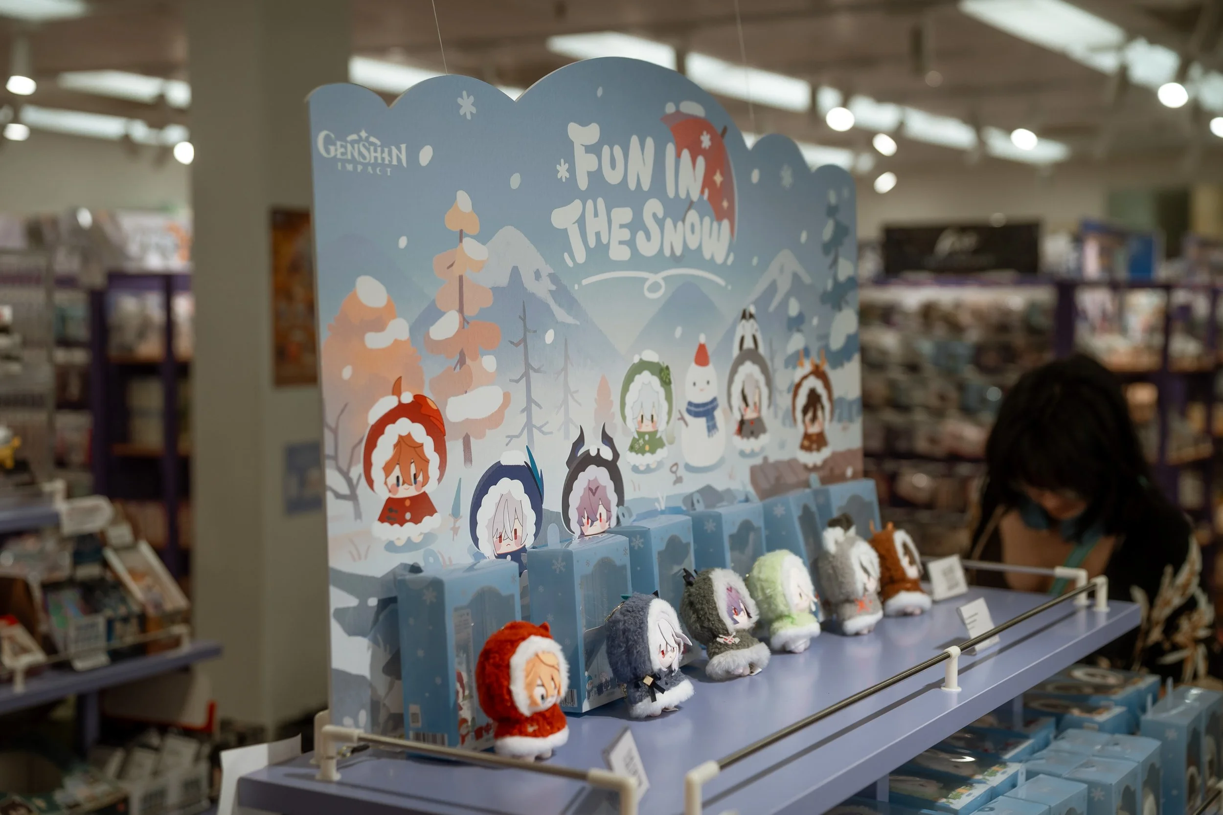 Display of plush toys and accessories themed for fun in the snow, with a sign that reads 'Fun in the Snow' and features cartoon characters in winter clothing amid a snowy mountain landscape at a store.