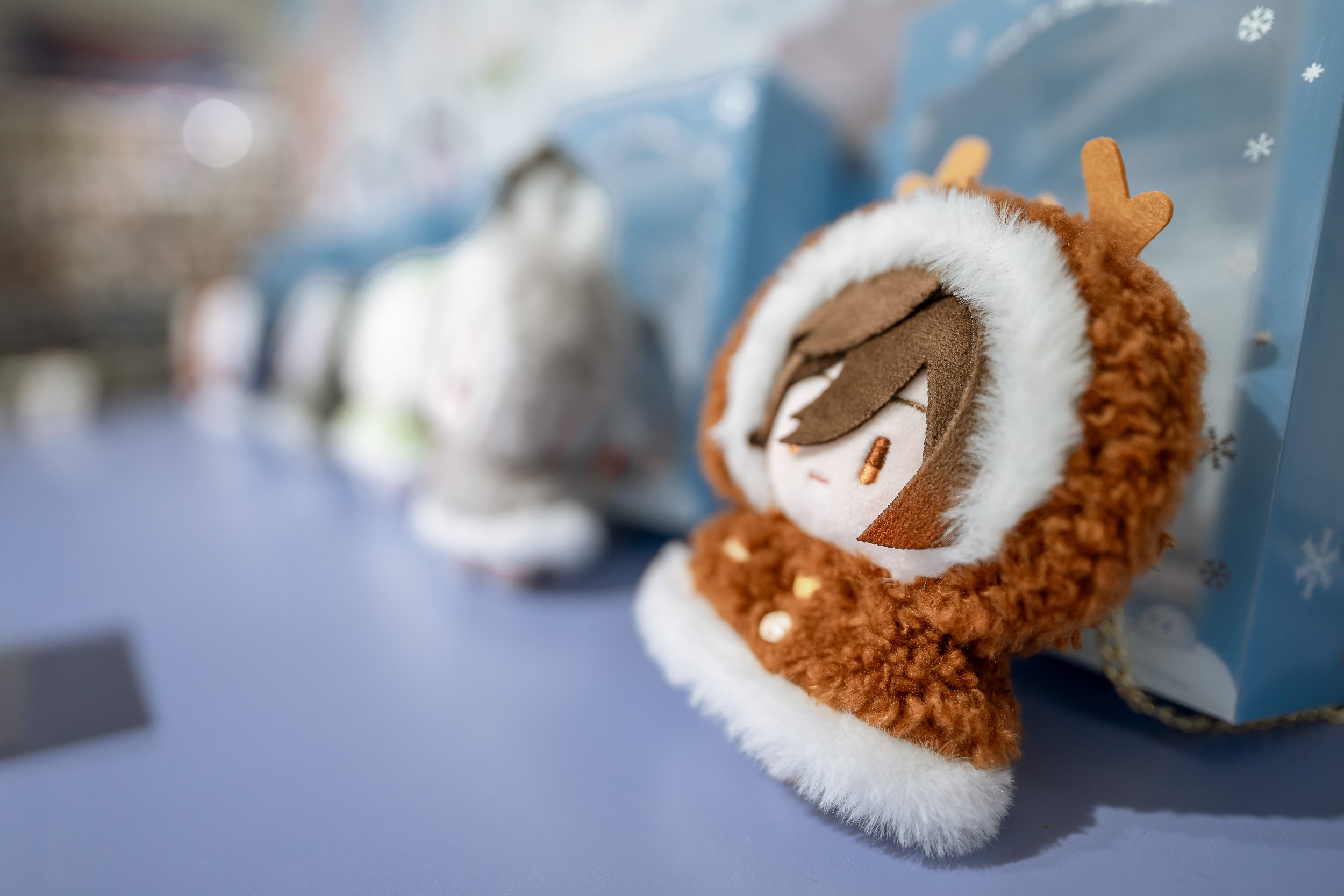 Close-up of a plush reindeer Christmas ornament with brown fur, white fluffy trim, and antlers, placed on a blue surface with a blurred background of other ornaments and winter-themed decorations.