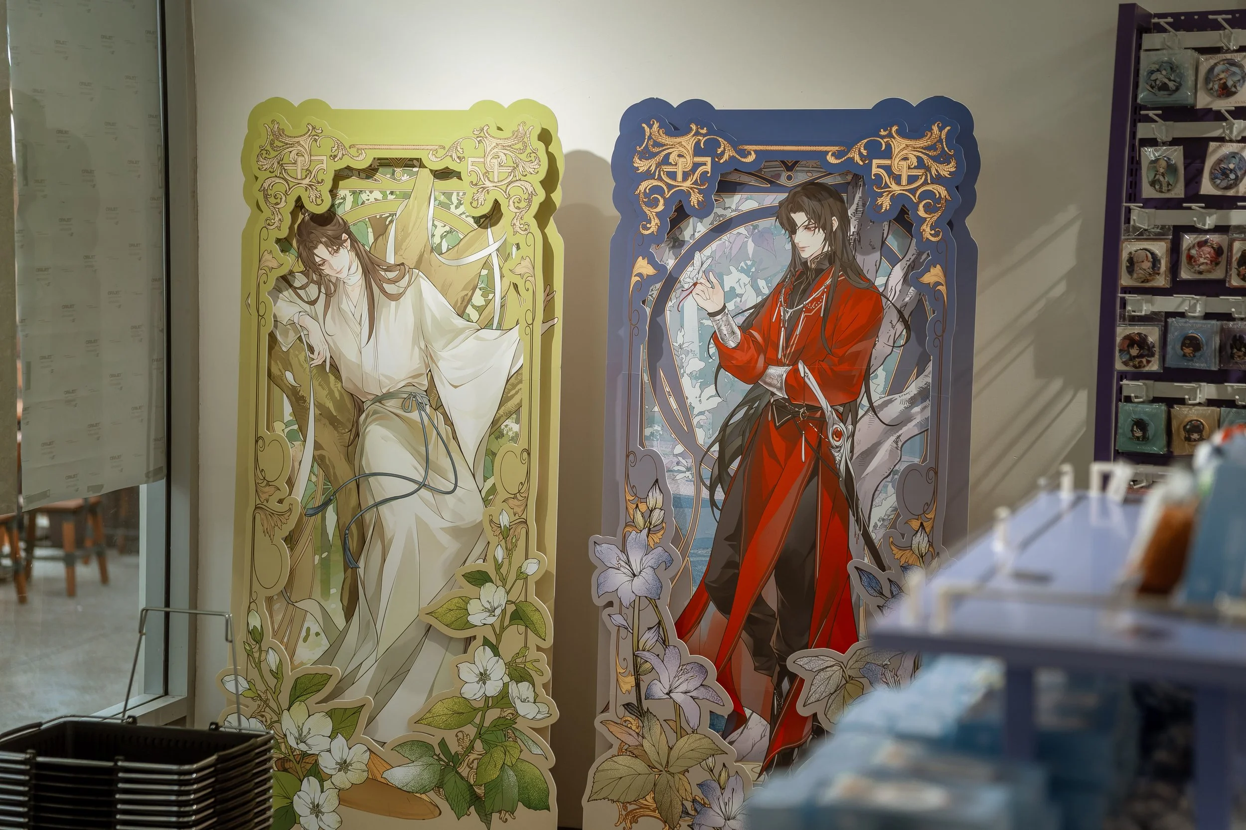 Two large paper cutouts with fantasy-style characters, one in white robes with long hair, the other in red robes with long dark hair, both framed with ornate gold and floral designs, displayed in a store.