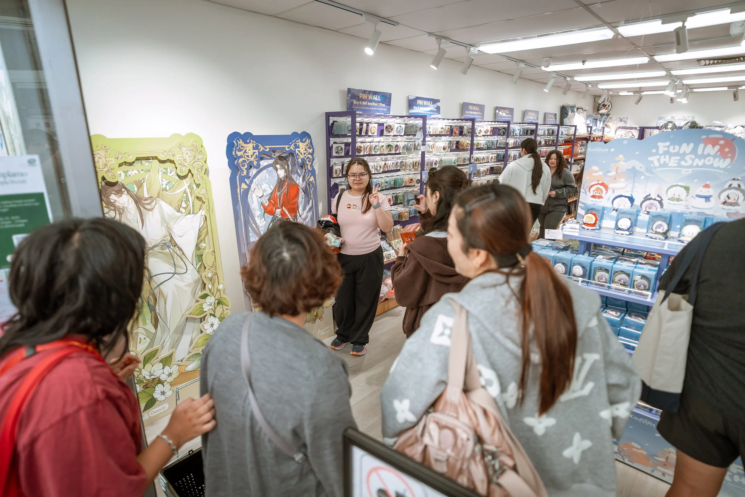 People shopping in a store with shelves stocked with various items, including anime or character-themed merchandise, some posters on the wall, and a group of shoppers, mostly women, looking at products.