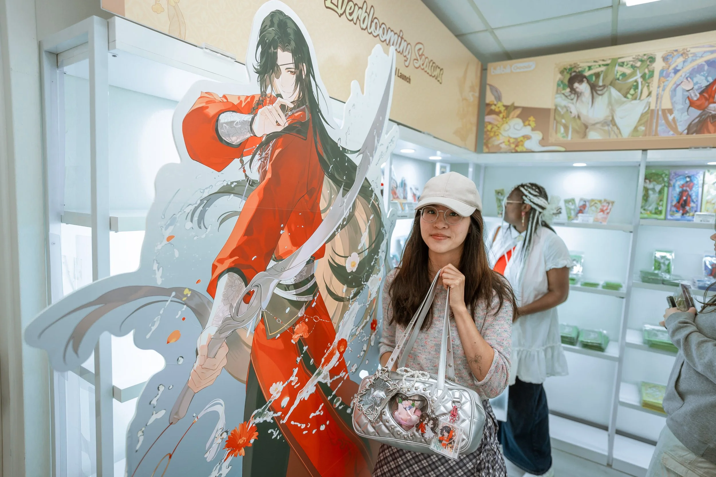 Woman with glasses, light-colored cap, and plaid skirt standing in an art exhibition, holding a silver handbag with anime-themed keychain, beside a large anime character cutout in a bookstore or gallery.