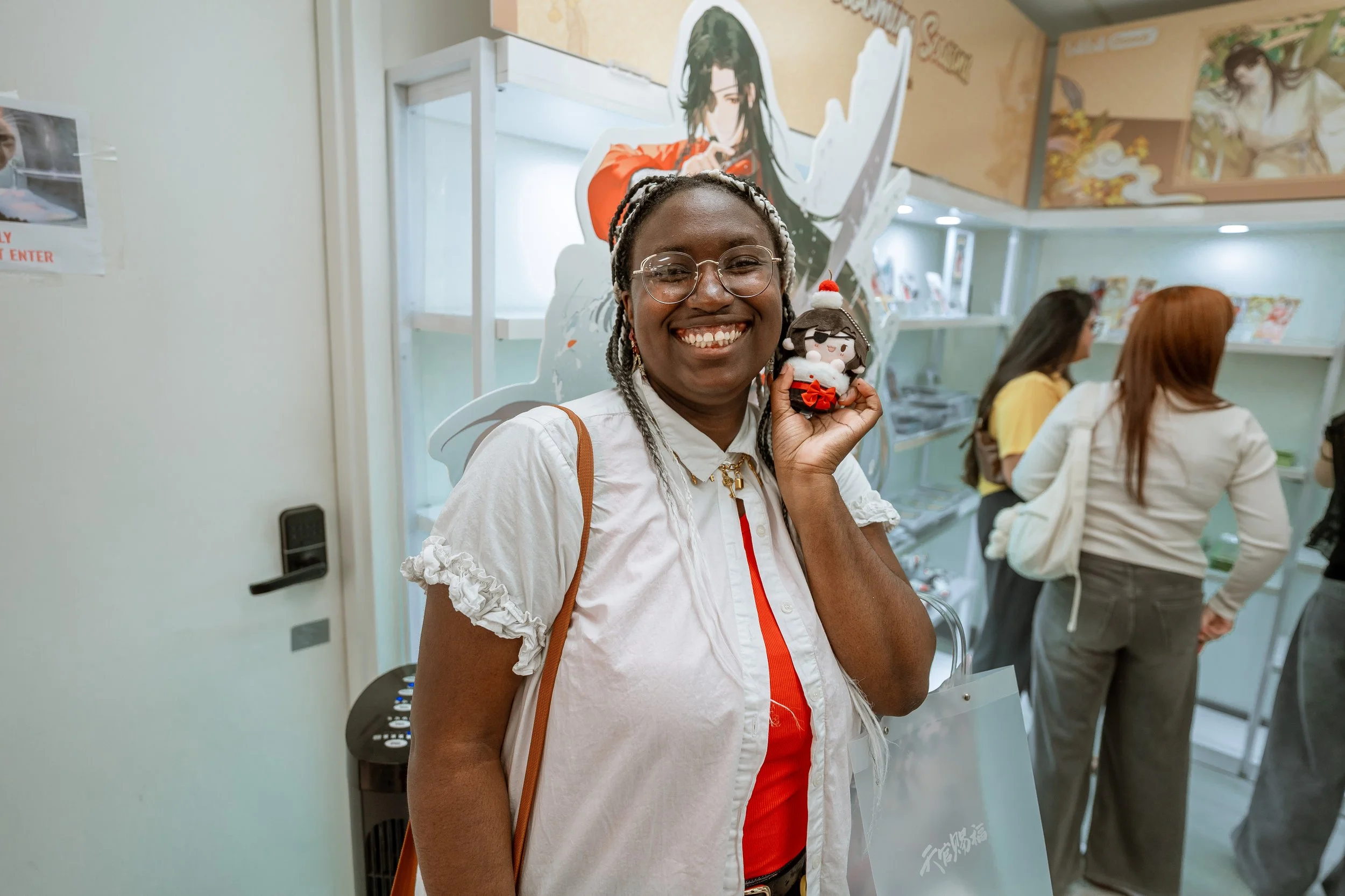 A smiling person with glasses and braided hair holding a small plush doll with a Christmas hat. In the background, there are three women sitting at a counter inside a store, with posters and shelves behind them.