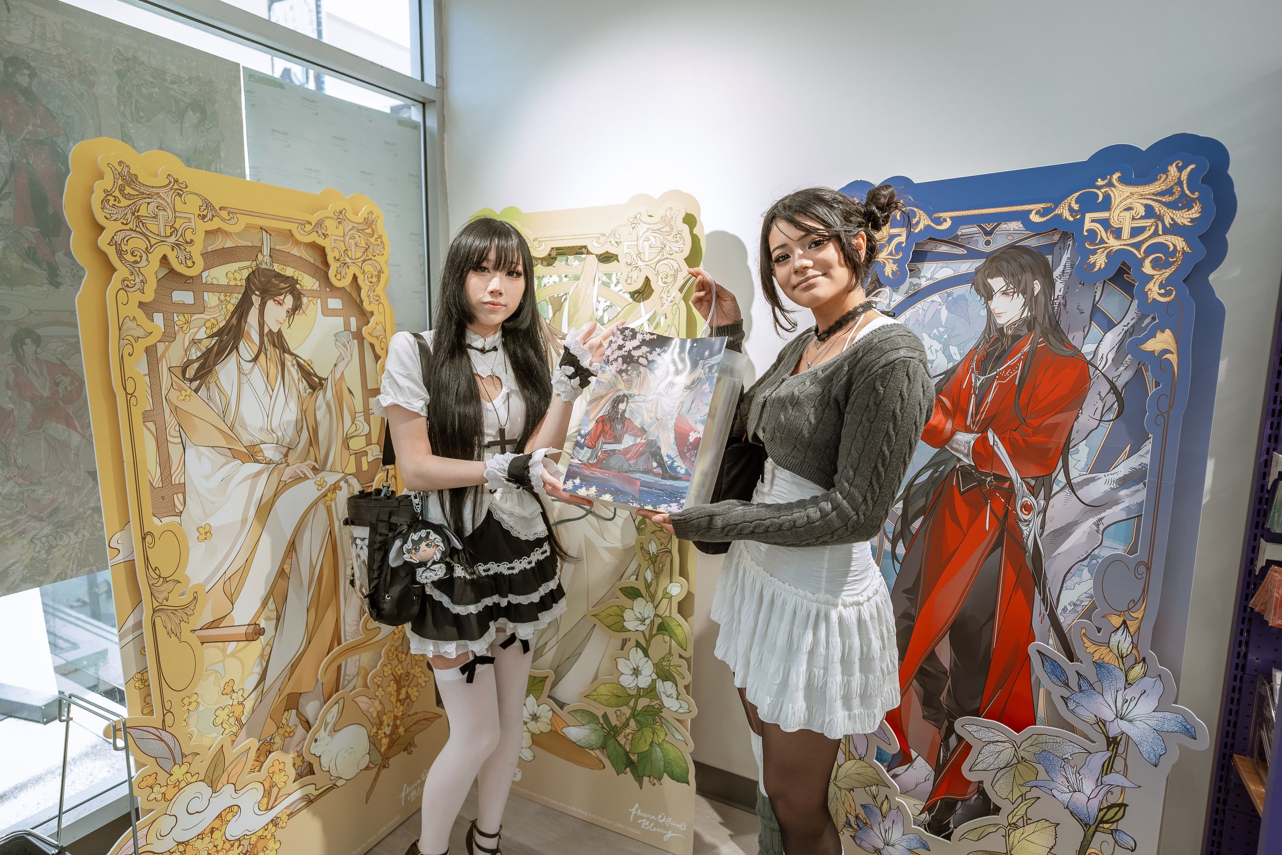Two women standing in front of colorful illustrated panels of fantasy characters at an art or exhibition display. One woman holds a bag with anime art, and both are looking at the camera.