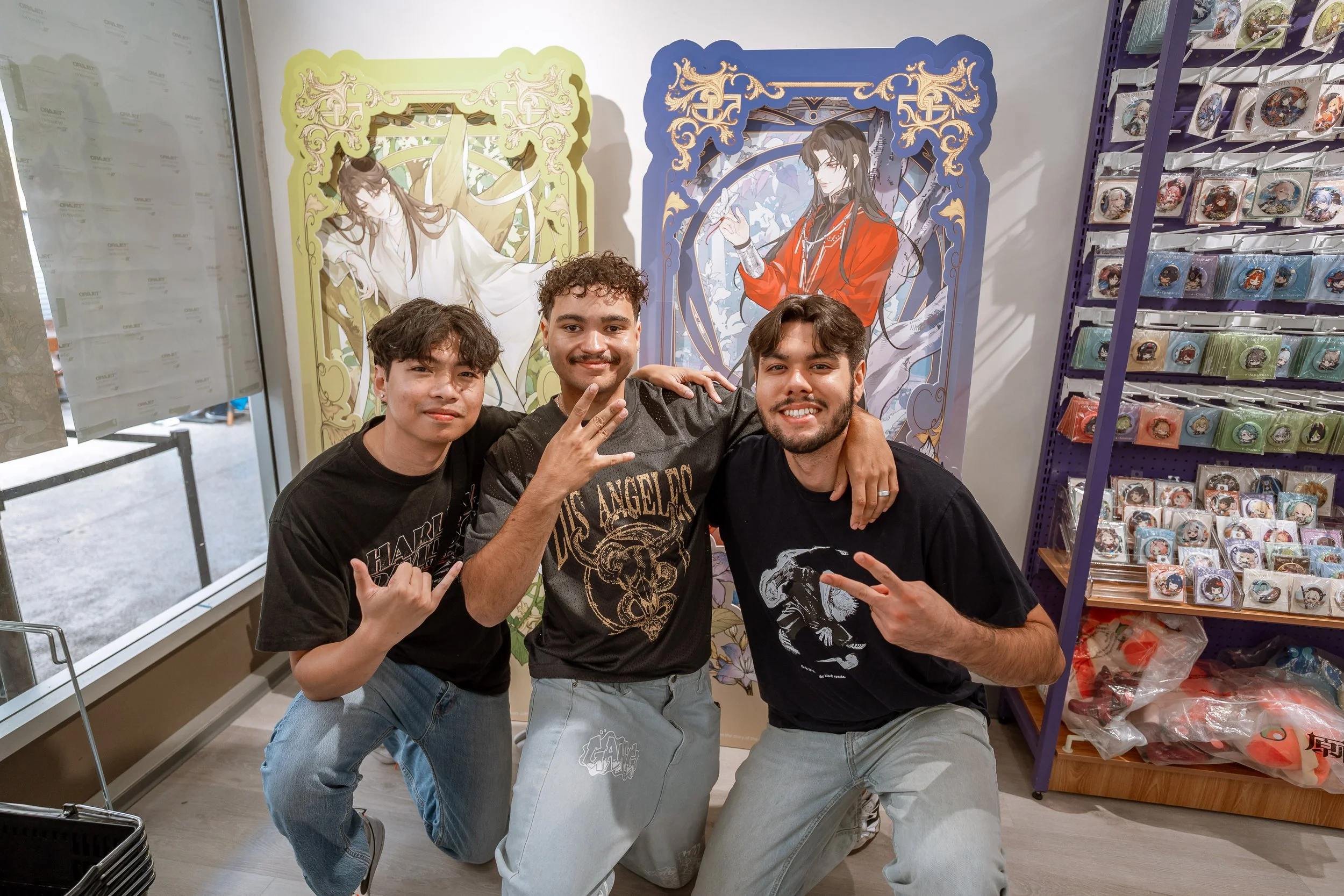 Three young men posing for a photo inside a store with anime posters and merchandise. They are smiling, and two of them are making hand gestures.