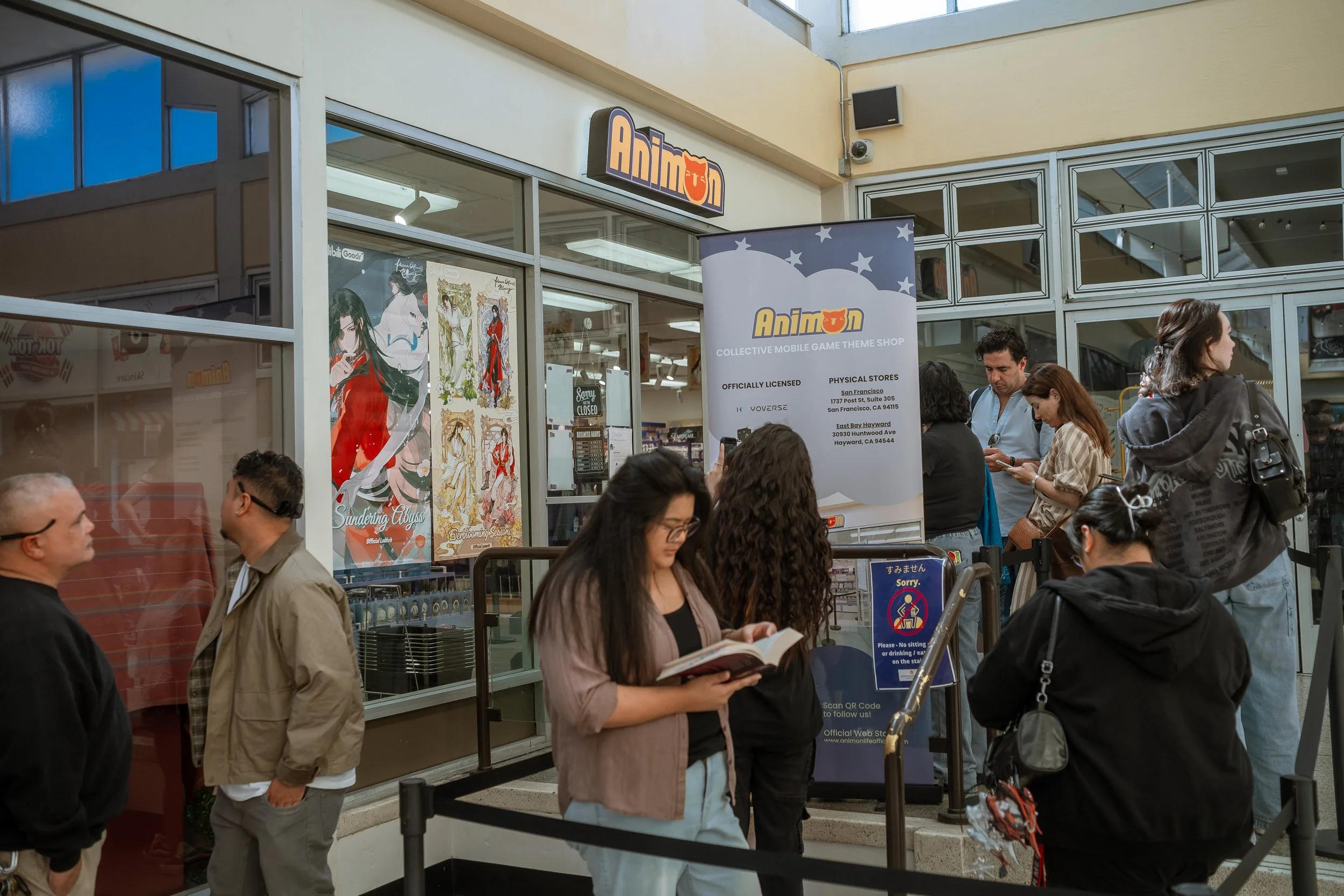 People inside and outside of an anime store named AnimOn, with posters of anime characters on the windows and a sign with store information. Several individuals are waiting in line or standing near the entrance.