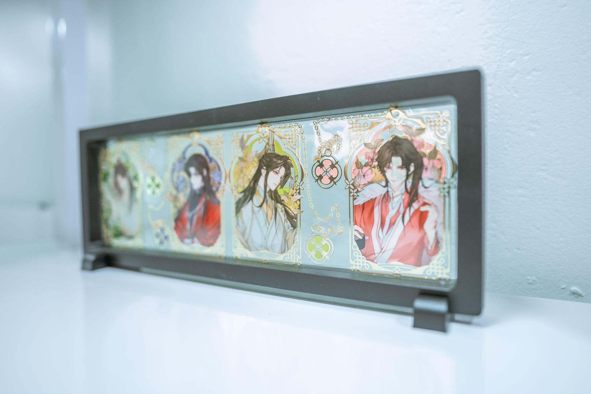 Close-up of a decorative printed paper inside a transparent plastic frame, featuring illustrations of two traditionally dressed Asian characters surrounded by intricate floral patterns with chains and ornate borders.