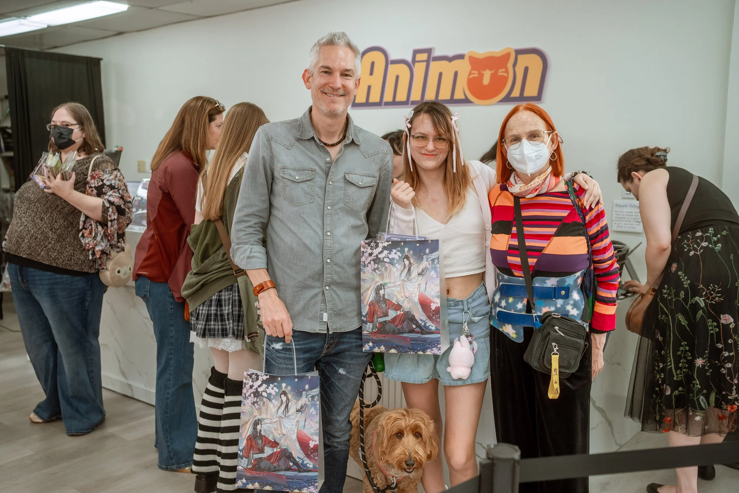 A group of people at an event, with a man and a woman at the center holding artwork, and a dog at their feet. They are standing in front of a light-colored wall with a sign that partially reads 'Anim'.