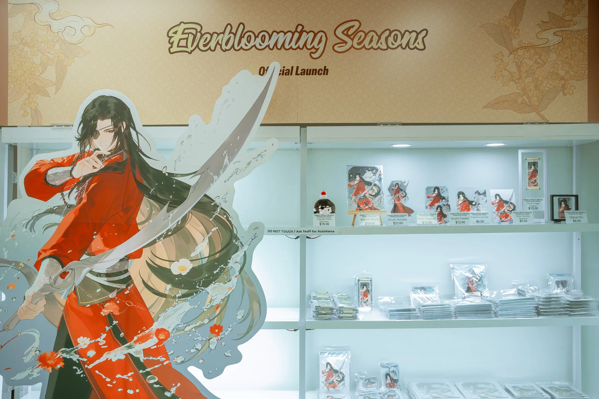 Display for the game 'Everblooming Seasons' at an event, featuring anime-style character art of a woman with long black hair in a red kimono with water splashes and flowers, with merchandise such as keychains, acrylic stands, and posters on shelves b