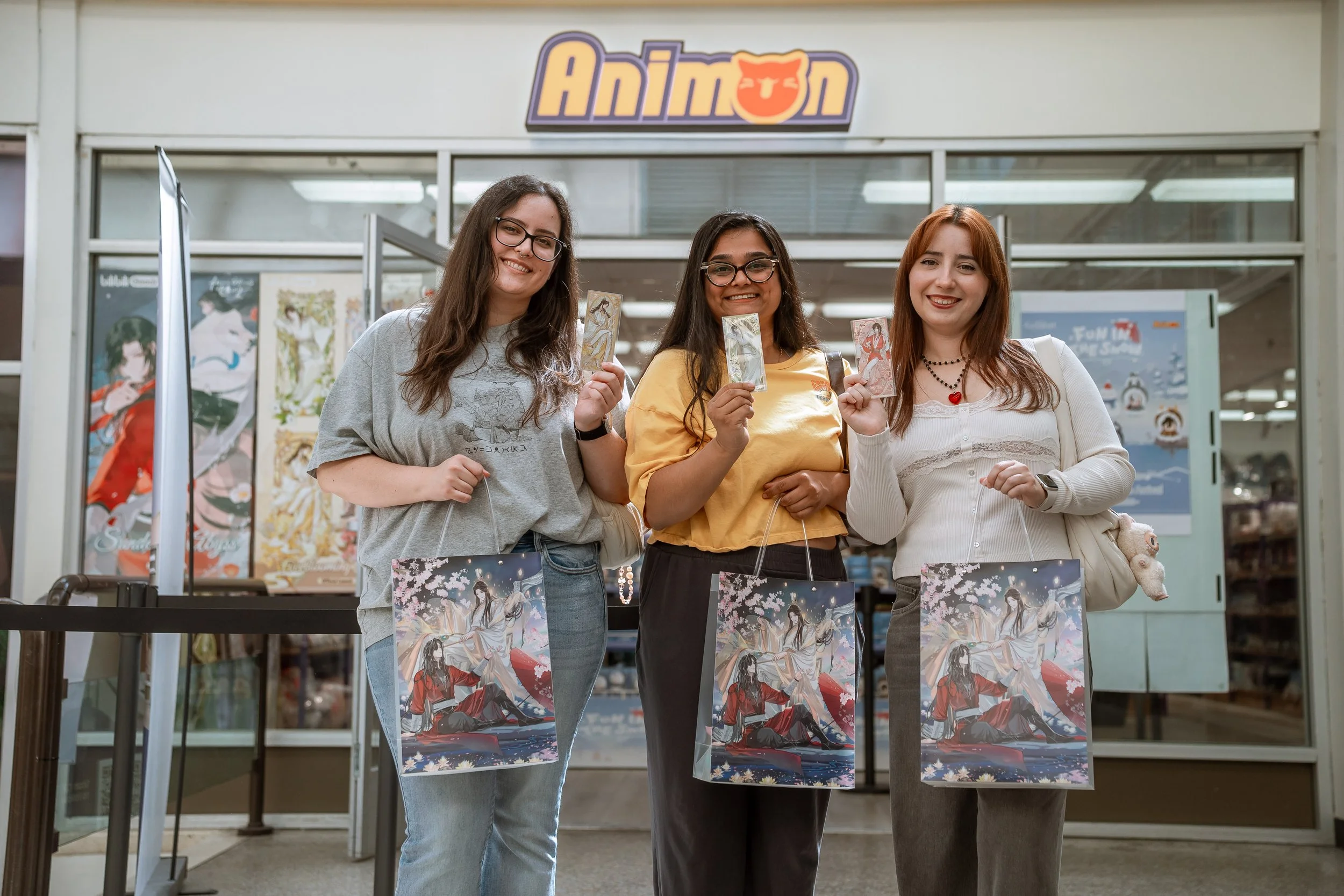 Three young women standing in front of an Anime store holding anime-themed posters and shopping bags, smiling at the camera.