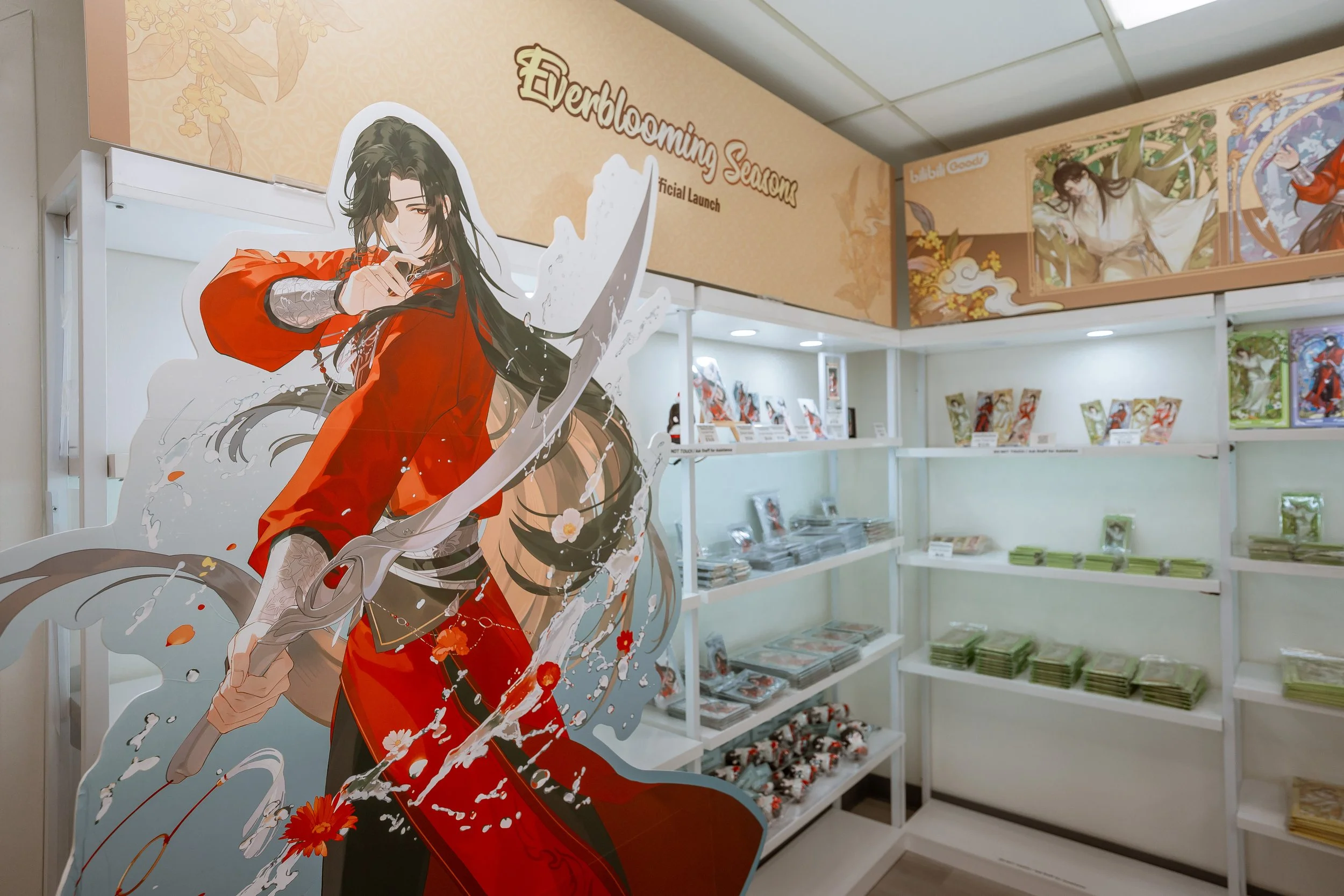 A store display for Everblooms Seasons featuring a large anime-style character cutout dressed in a red kimono with floral patterns. The character has long dark hair and is holding a sword. The background includes shelves with books, manga, or art pri
