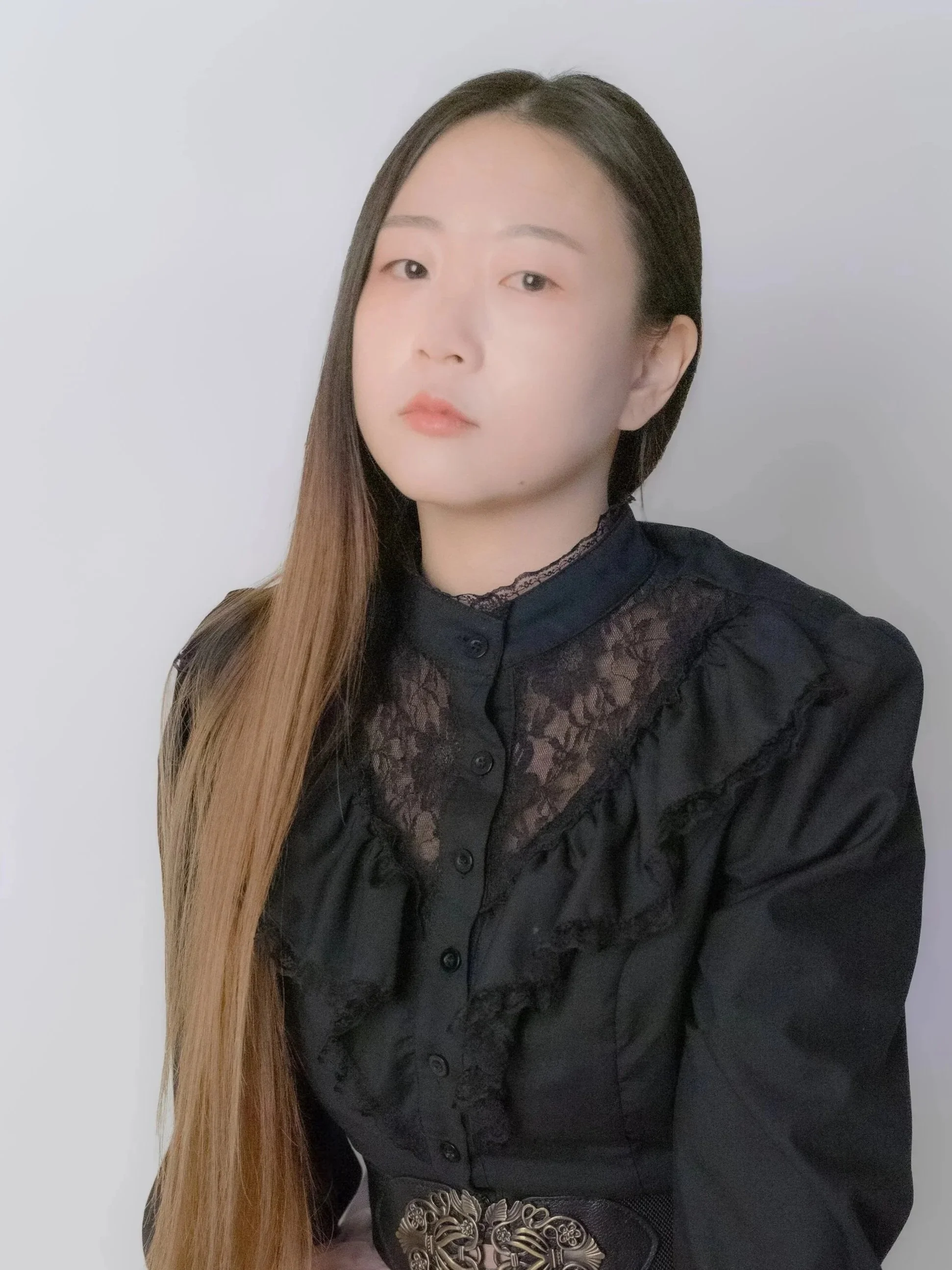 A woman with long brown hair wearing a black lace blouse with a decorative belt, looking at the camera with a neutral expression, standing against a plain white background.
