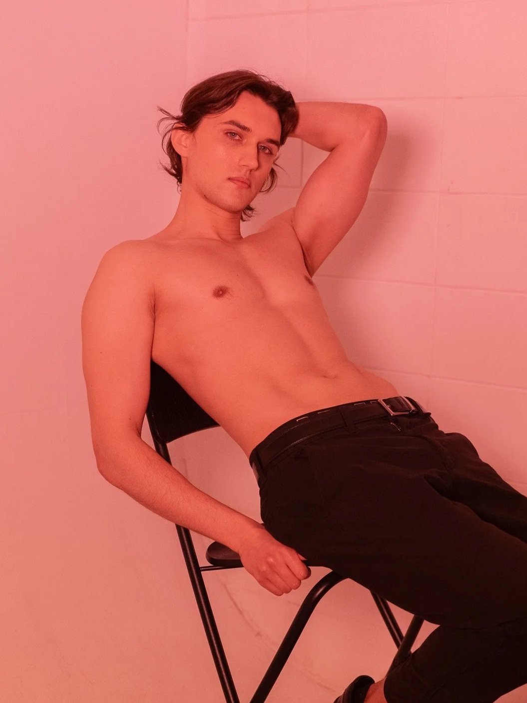 A young man with short brown hair and fair skin, sitting shirtless on a black folding chair, with a relaxed pose, one hand resting behind his head, looking at the camera, against a plain light-colored wall, with a pinkish hue lighting.