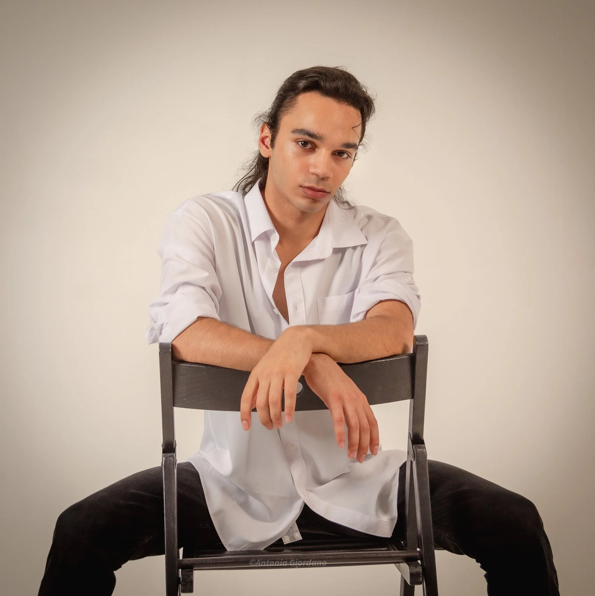 Young man with long dark hair, wearing a white shirt, sitting on a chair backwards with arms resting on the top of the chair, against a plain beige background.