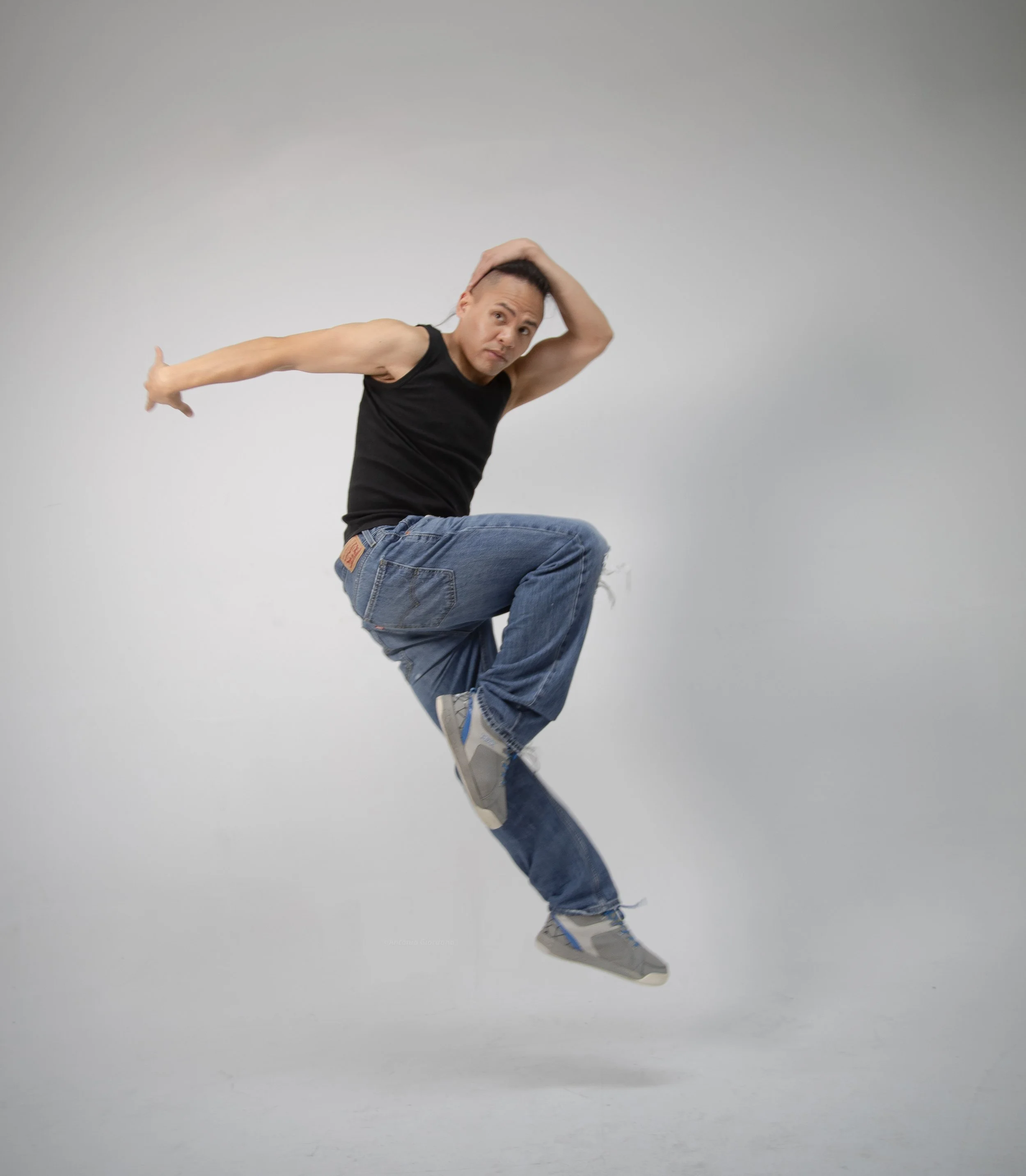 A man wearing a black tank top, jeans, and sneakers is mid-air, jumping with one hand on his head and the other arm extended outward against a plain white background.