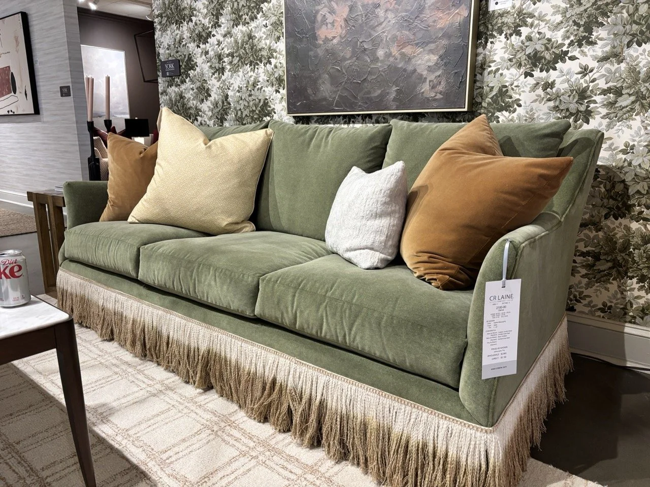a soft green sofa with fringe detail and golden throw pillows against a wall with leaf patterned wallpaper seen by cat french design at high point furniture market spring 2026 home design trends