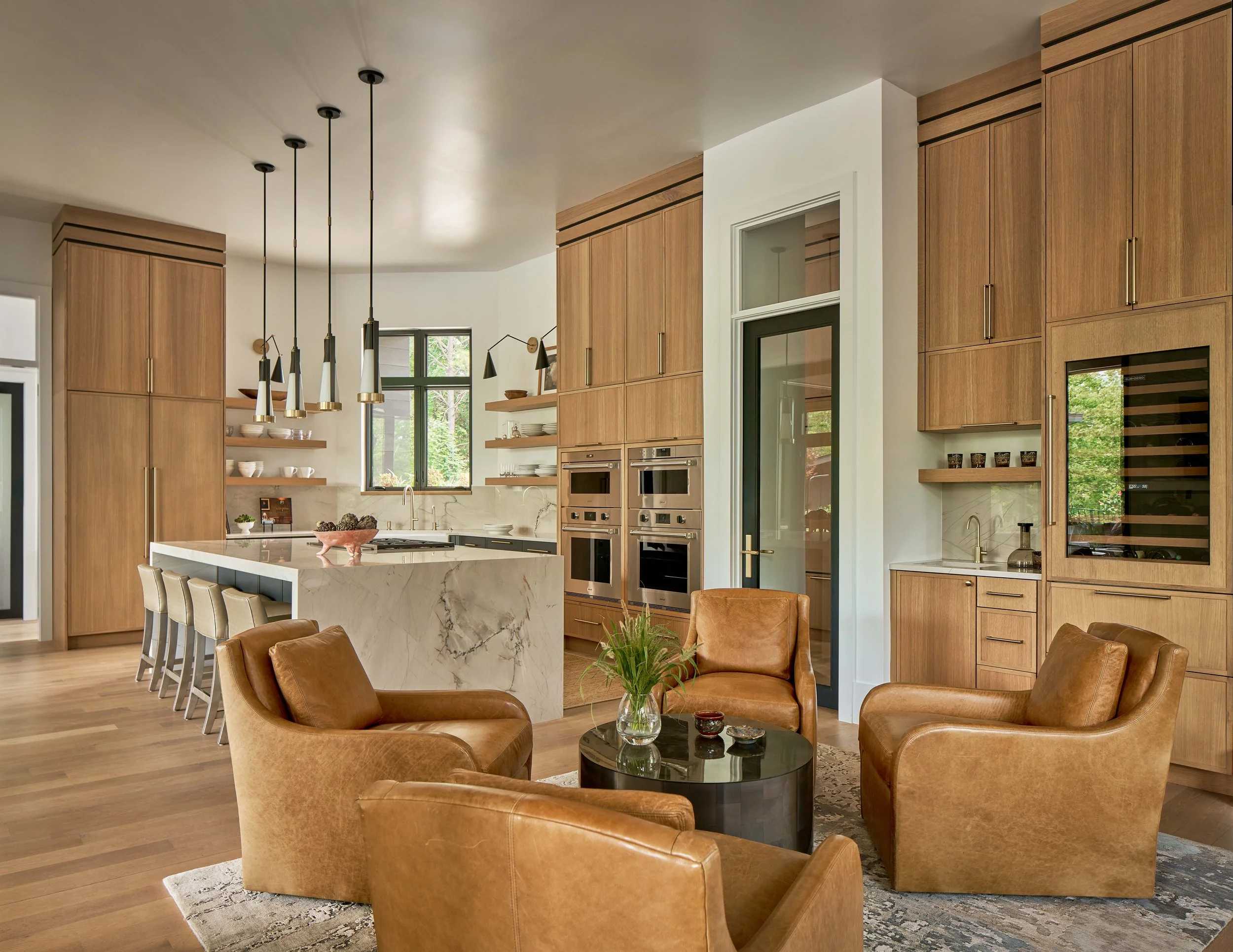 leather swivel armchairs and custom built-in bar with wine fridge and sink next to a custom kitchen with large quartzite waterfall island in a durham custom home design by cat french design interior designer for custom home warm luxury interiors