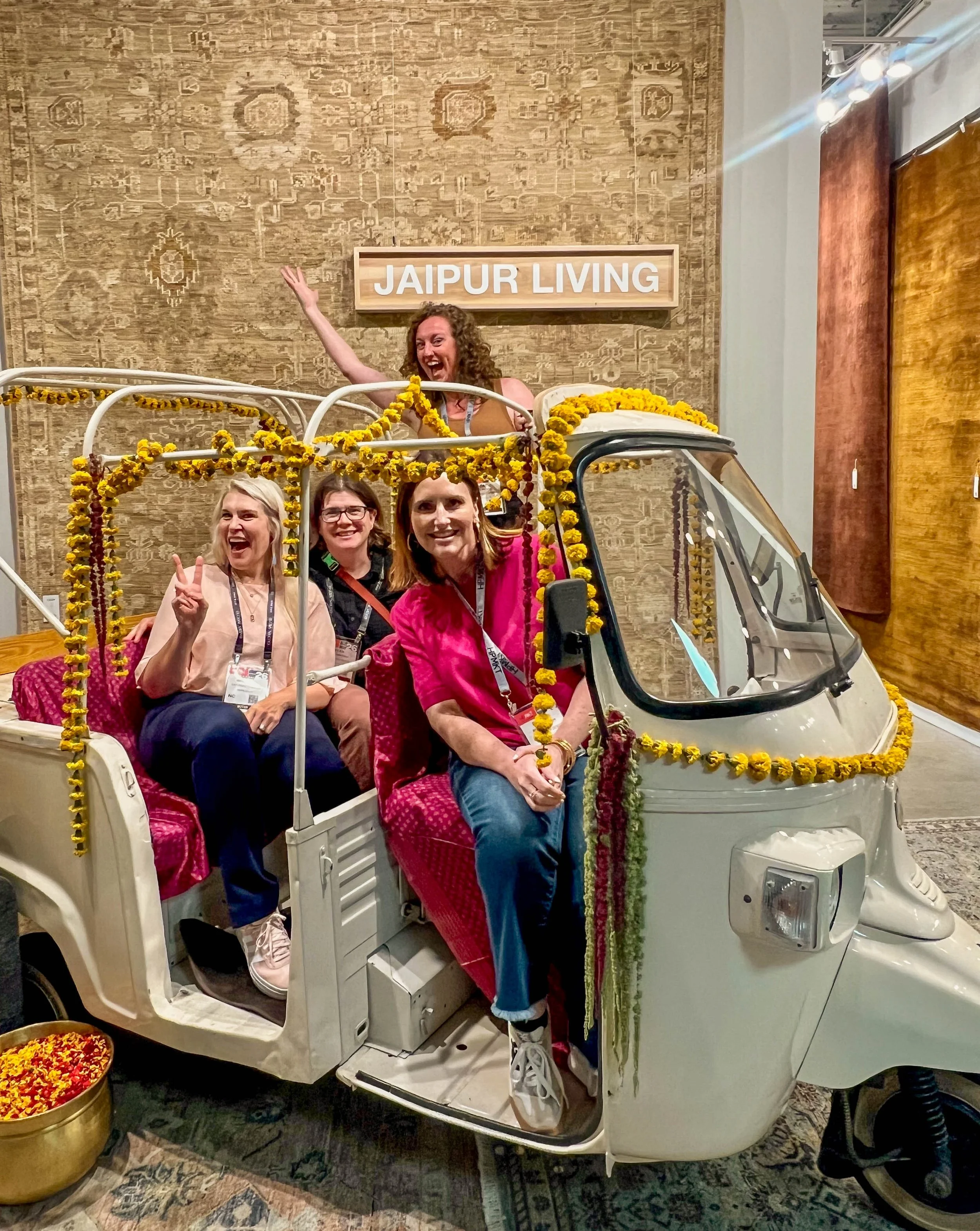 four women in a vintage auto rickshaw decorated with dried flowers underneath a large rug hanging on the wall and a sign that says jaipur living at the high point furniture market 2026 jaipur showroom