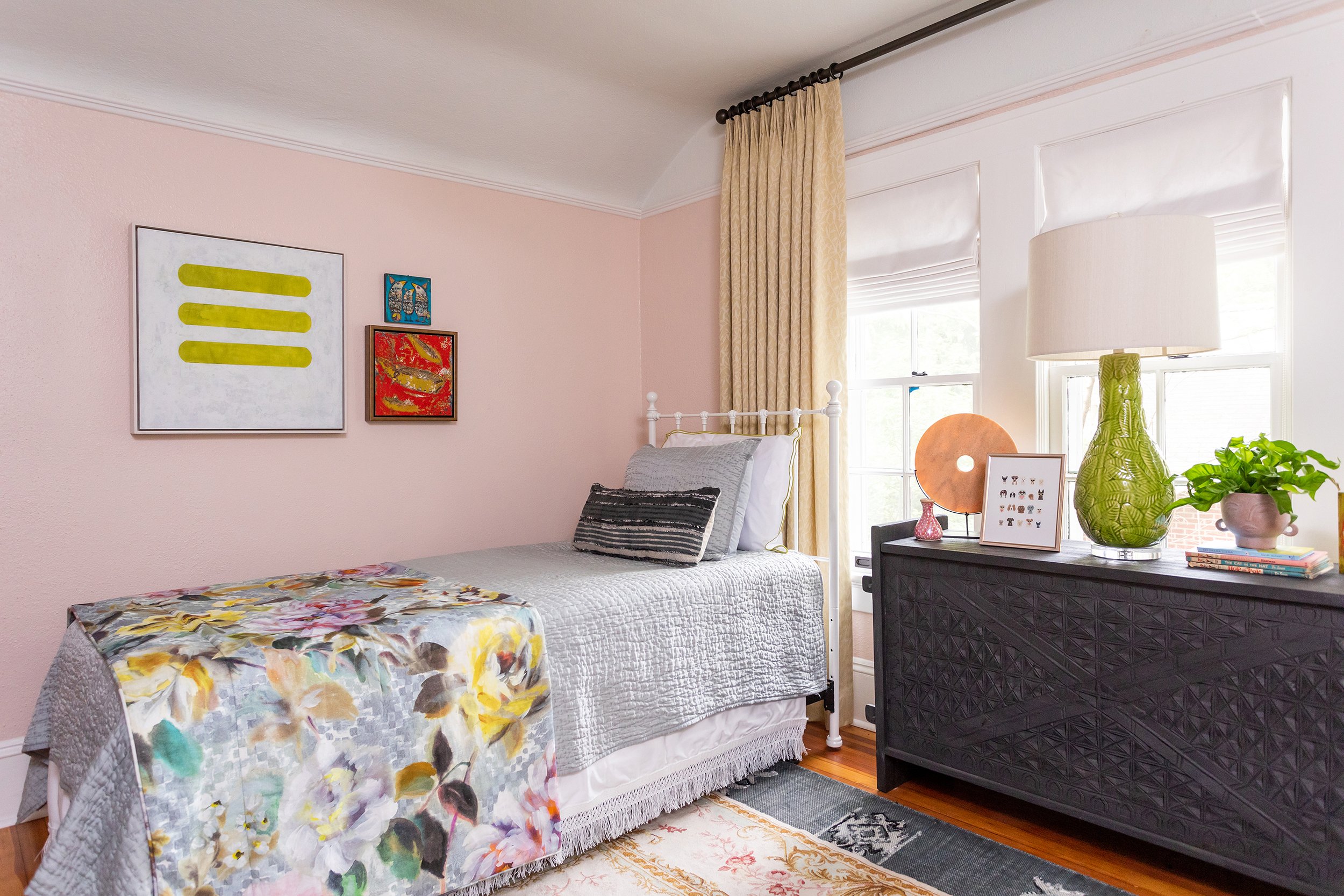a guest room with warm pink walls and a white twin bed with floral bedspread and black console with green lamp designed by cat french design durham interior design chapel hill interior design