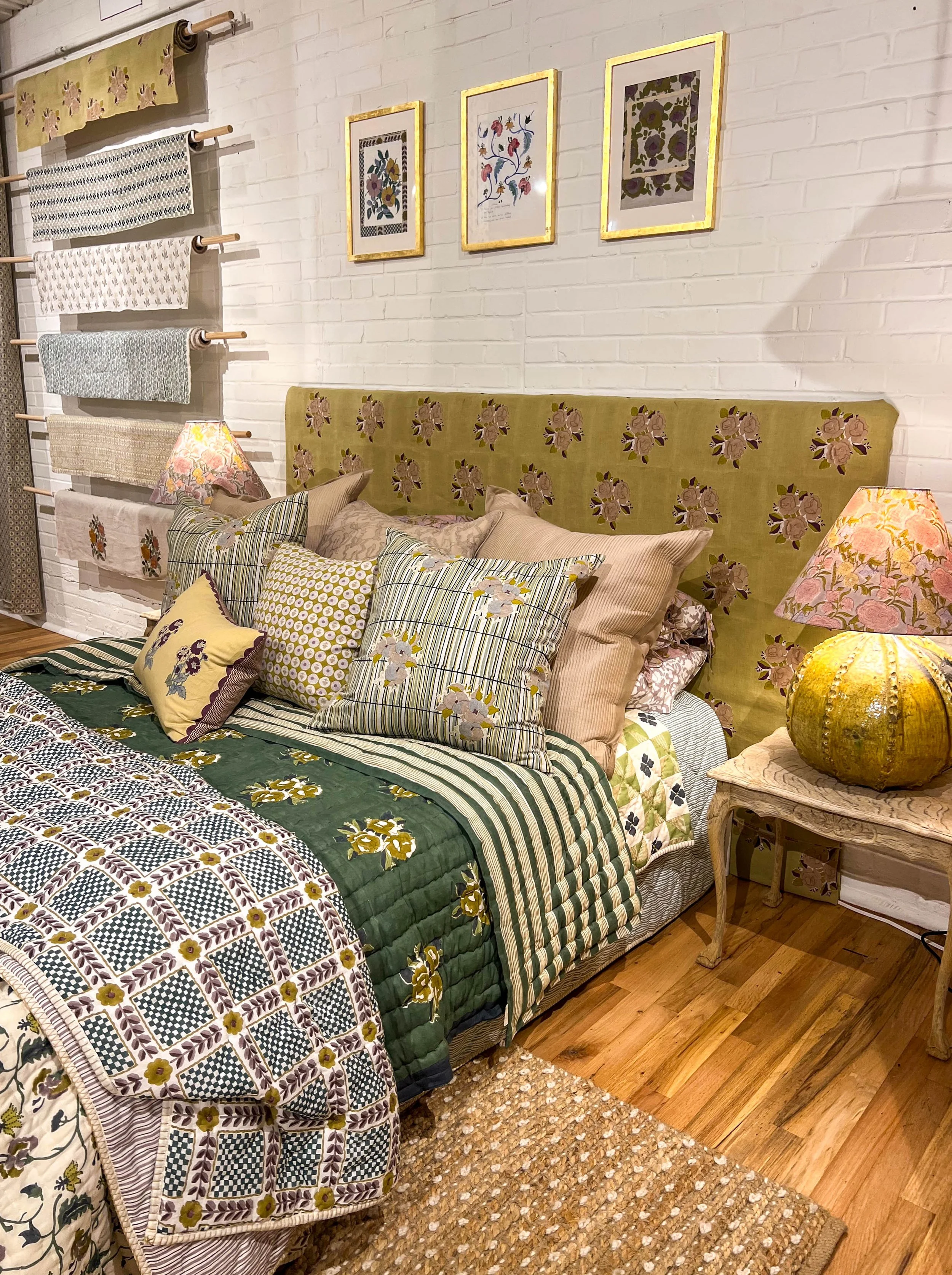 a bedroom scene in a showroom with a bed made up with handcrafted linens in floral and nature-inspired patterns an upholstered headboard with green fabric and pink flowers and two bedside lamps with floral lampshades