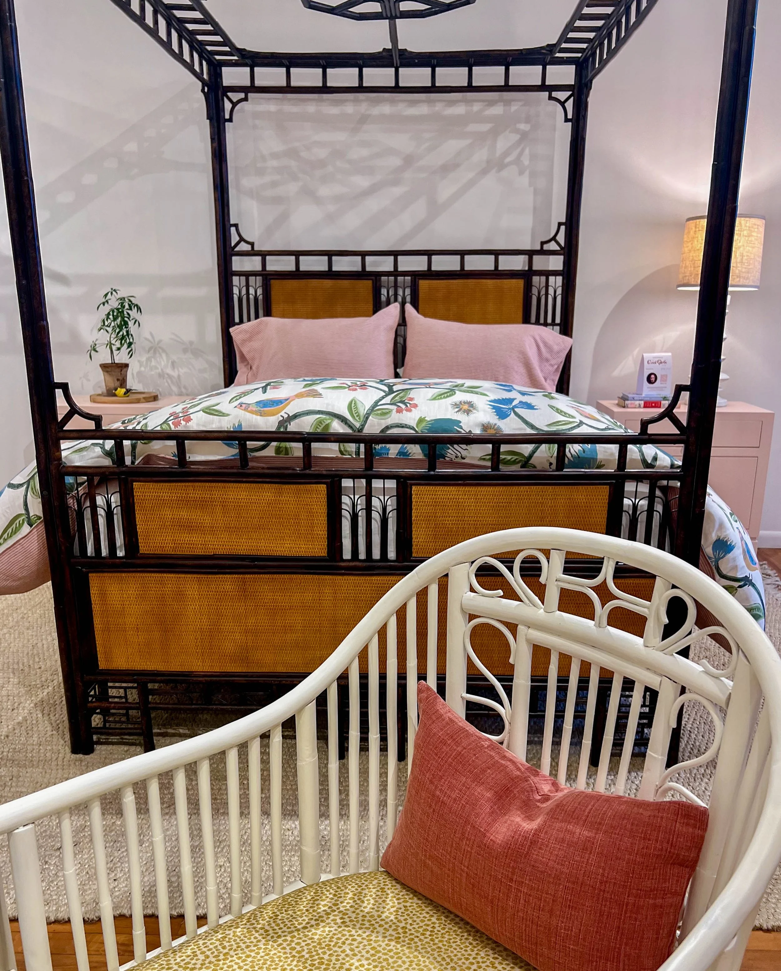 a soft white colored cane armchair with yellow cushion and red throw pillow in the foreground with a black and burnished wood cane bed eclectic style with a floral coverlet and light pink bedside tables in a showroom at high point furniture market