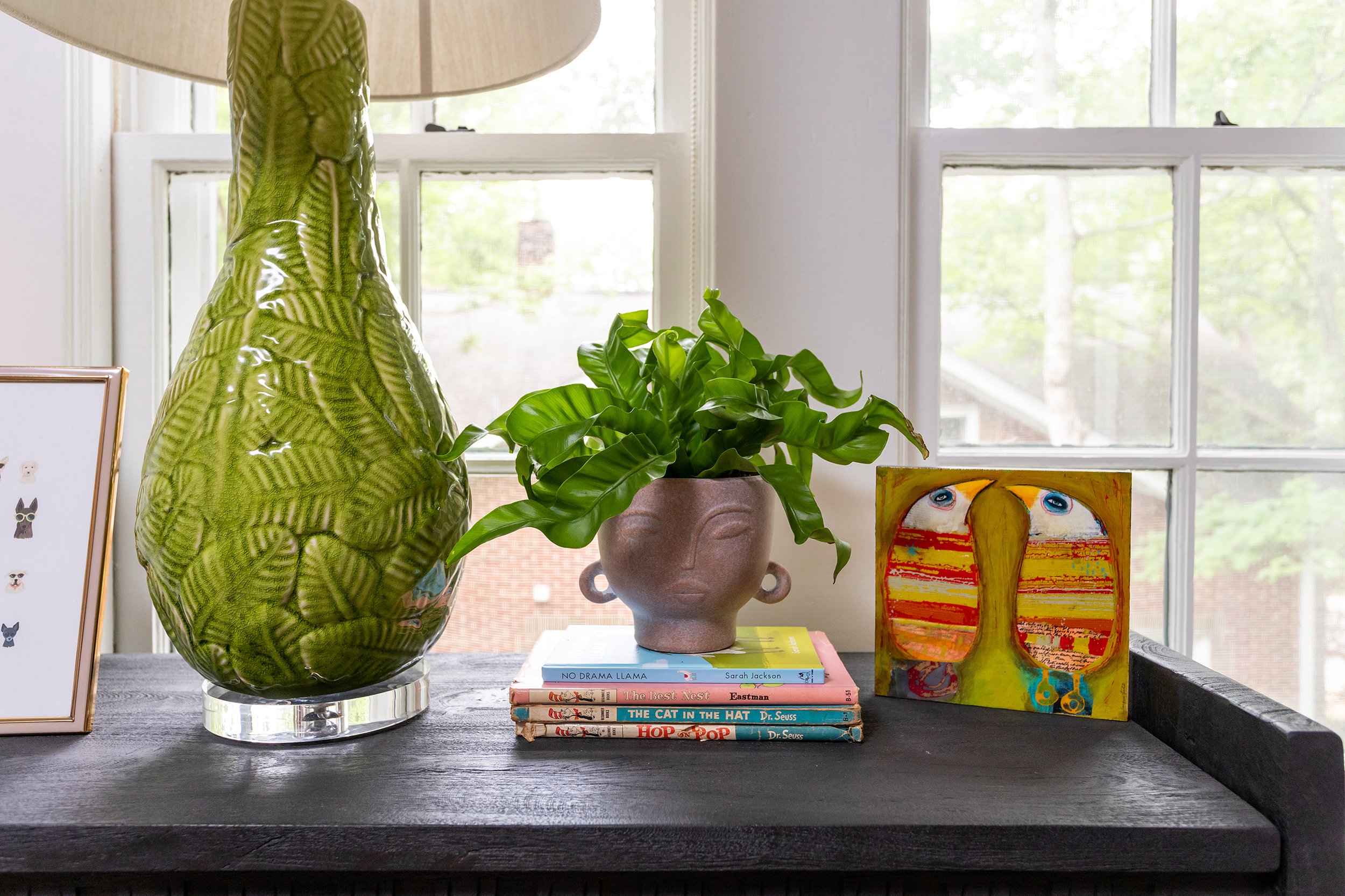 a black bedside table with textured green lamp house plant and colorful bird art in a guest room designed by cat french design award winning interior designer durham interior designer