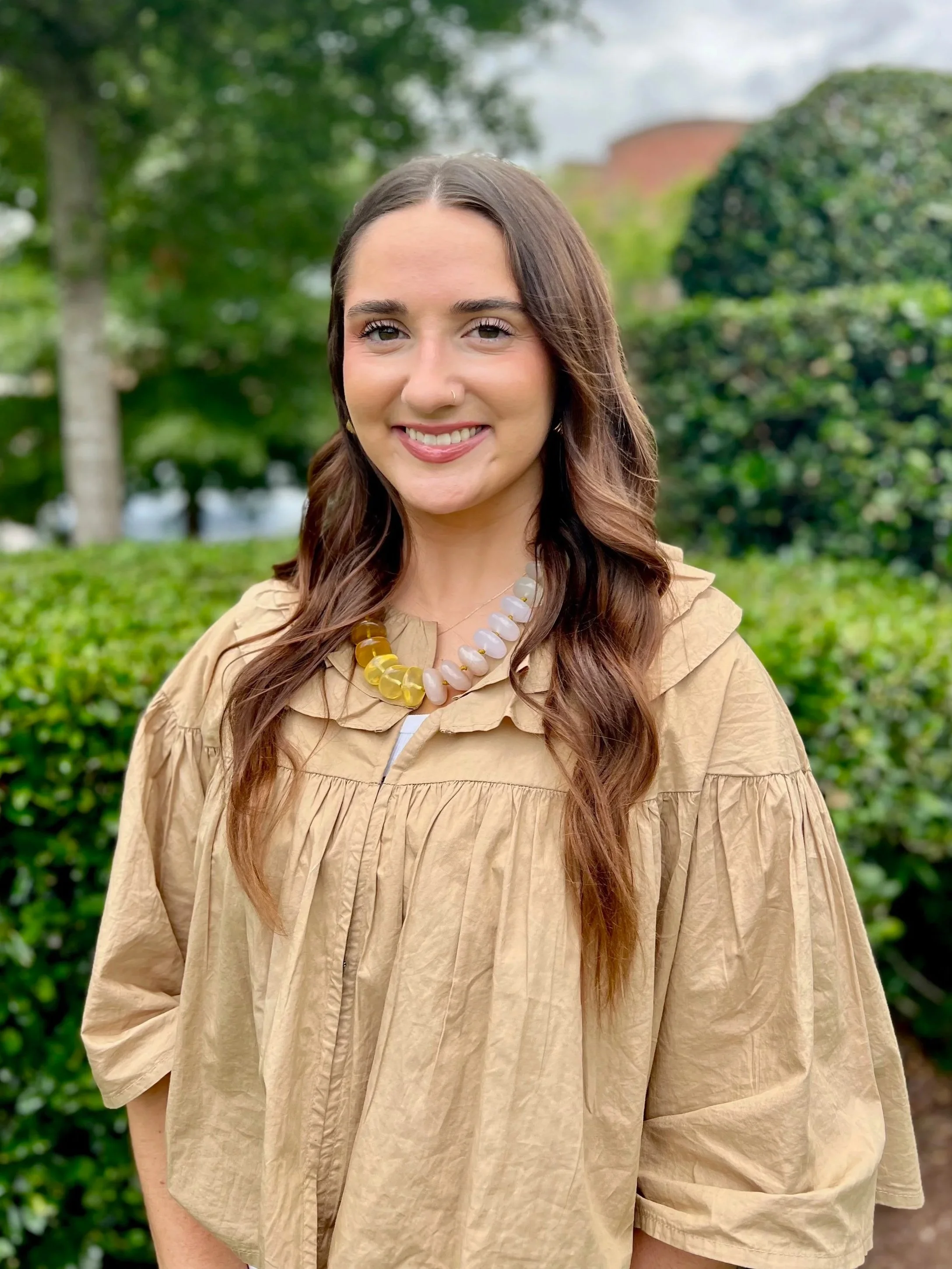Smiling woman with long dark hair wearing a beige blouse, standing outdoors near a hedge with trees and sunlight in the background.