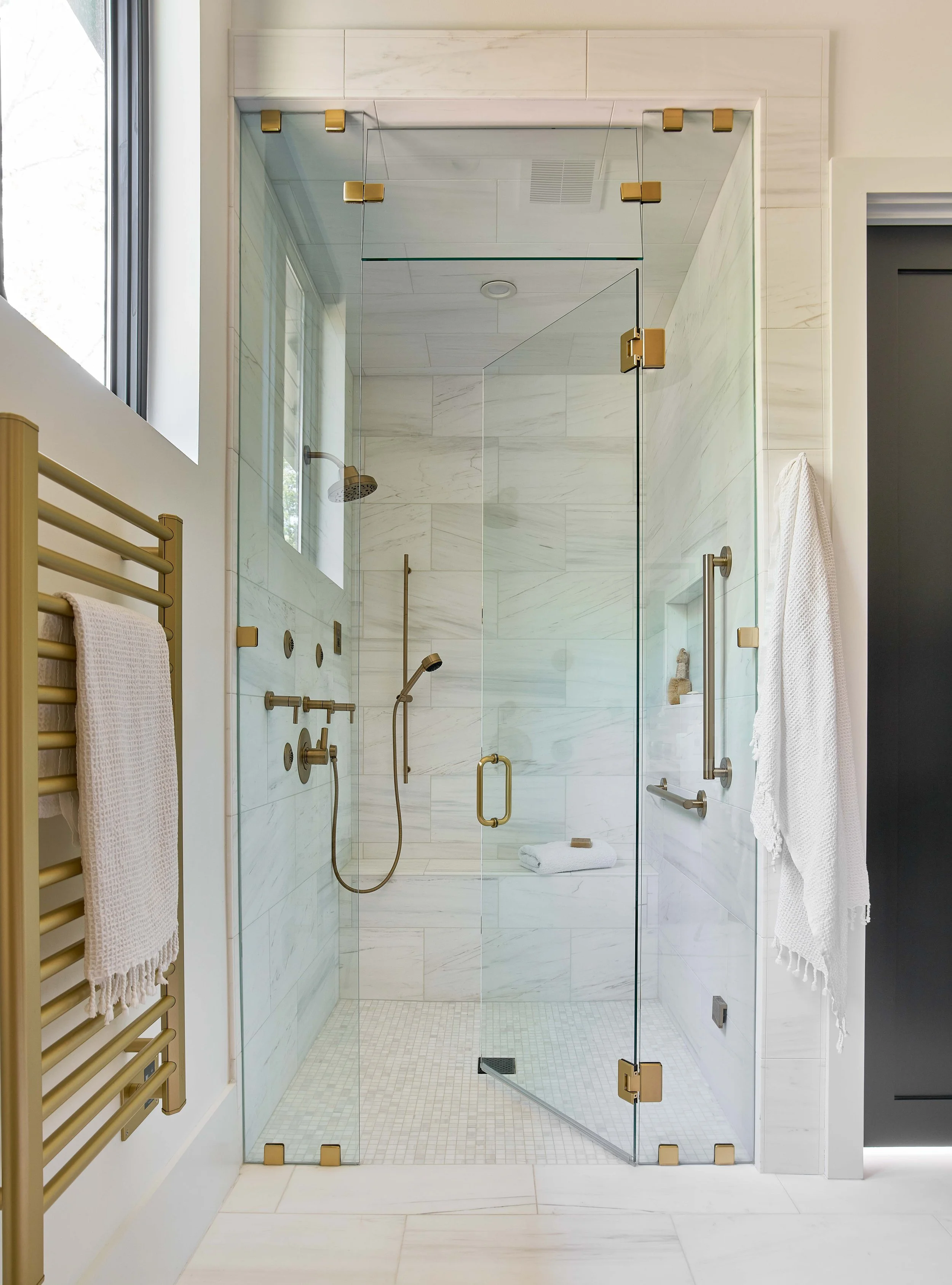 spa shower with gold hardware and glass shower door white and gray tile with built-in shower bench and a modern gold heated towel rack in a dual bathroom primary suite durham north carolina custom home aging in place home design zero entry shower
