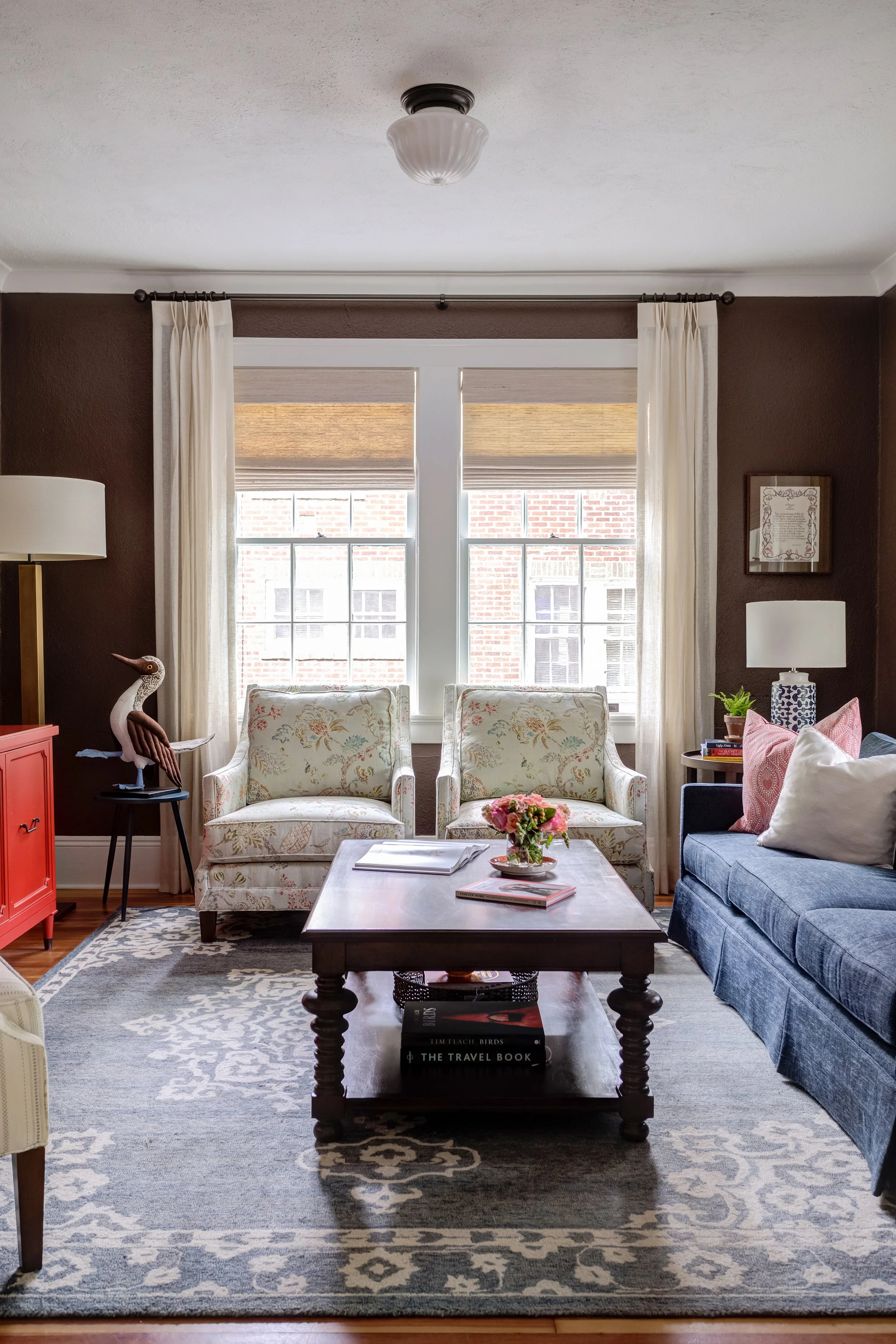 deep chocolate wall color in a den with large window, floral armchairs blue couch blue rug and wood coffee table designed by cat french design chapel hill interior designer durham interior designer