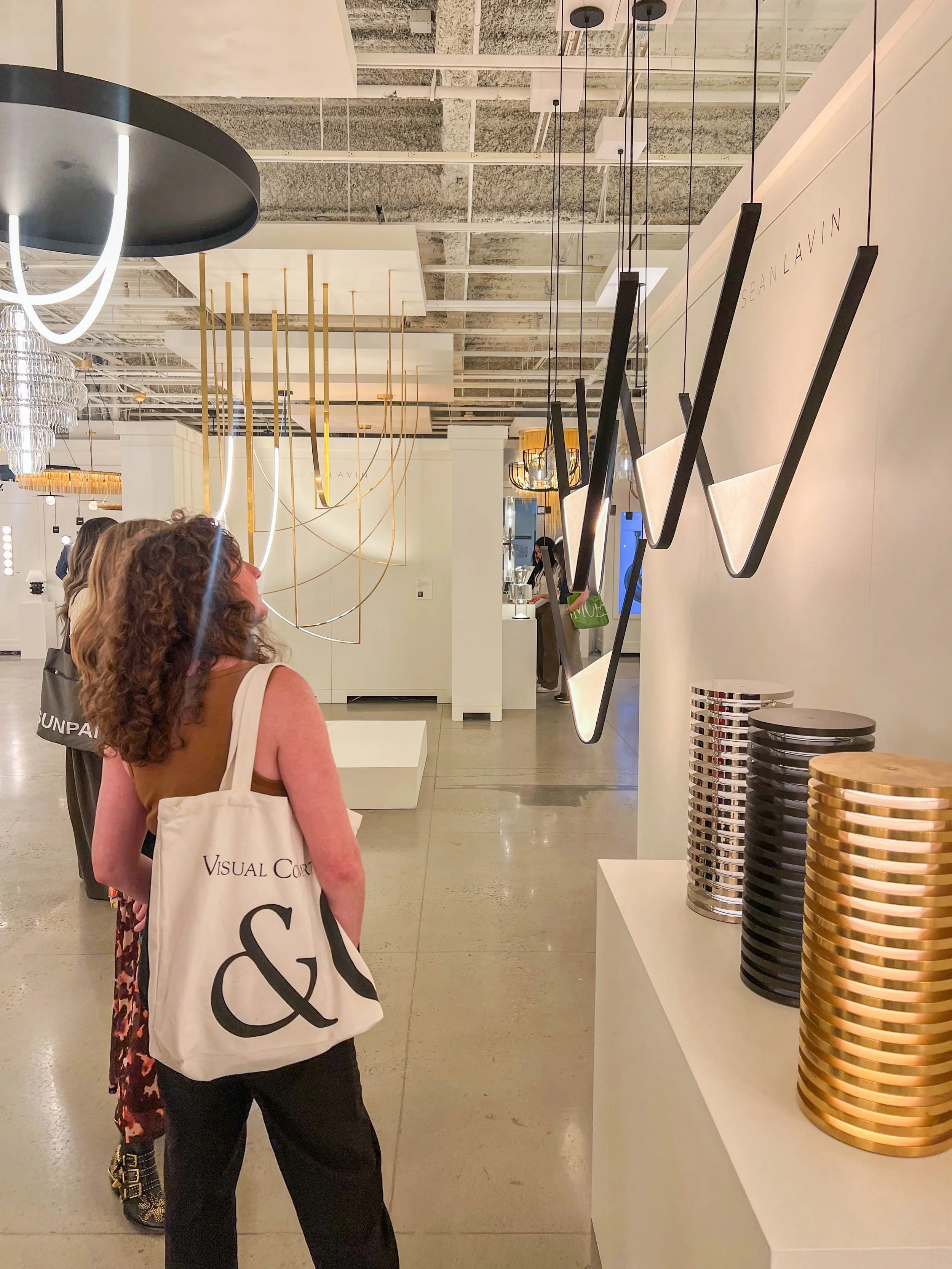 woman wearing black pants and brown shirt with curly hair carrying a canvas tote bag in a lighting showroom viewing sculptural metallic finish lamps and chandeliers at high point furniture market spring 2026